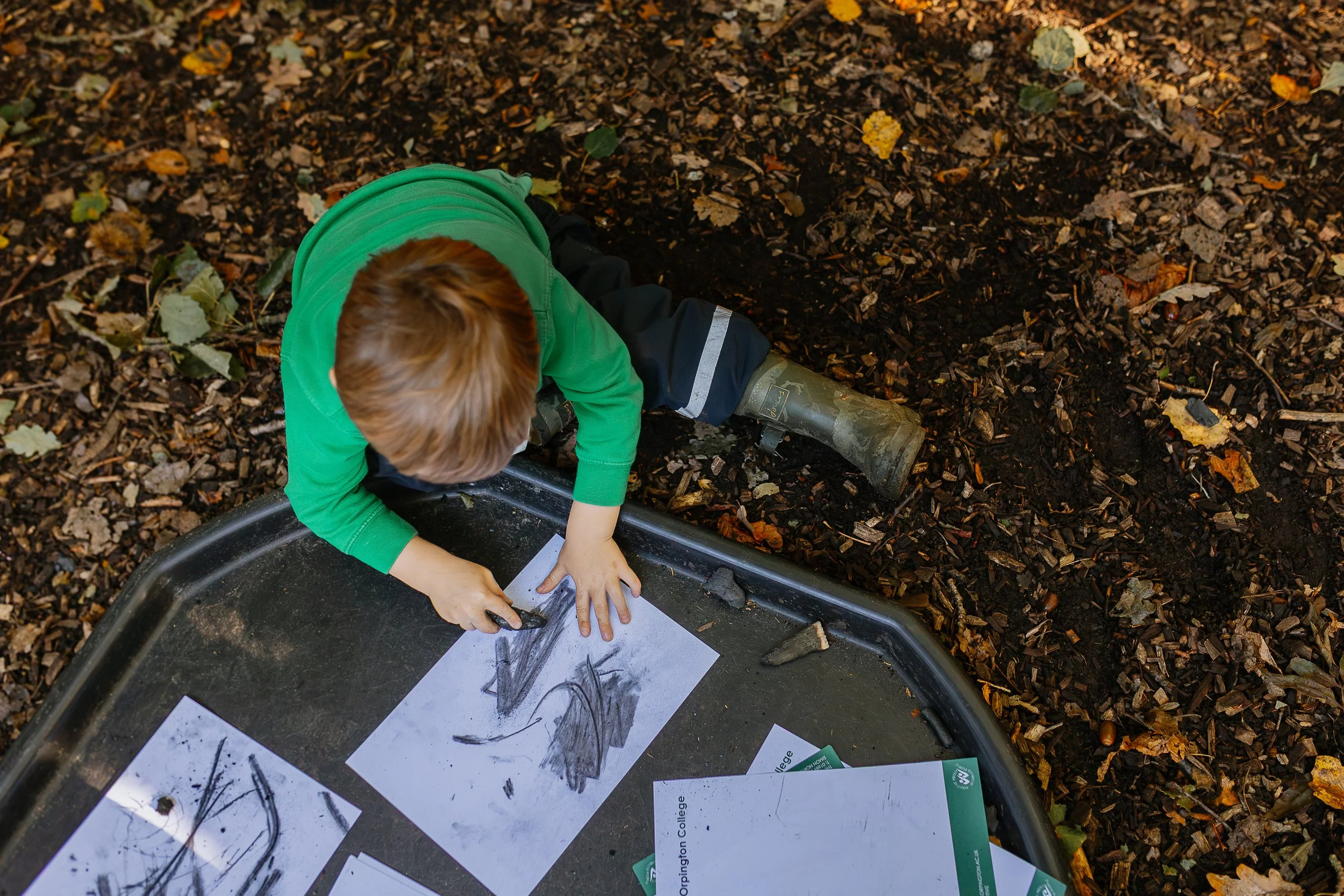 A young boy with red hair wearing a green shirt and rain boots is drawing on a paper with black marker, positioned near a shallow planter with paper and drawings inside, surrounded by a leafy wood chip ground.