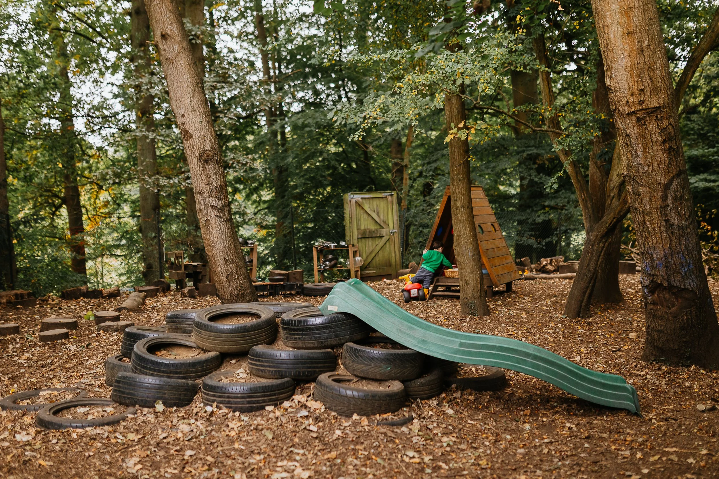 A wooded outdoor play area for children with tires, a slide, a wooden house, and a toddler playing.