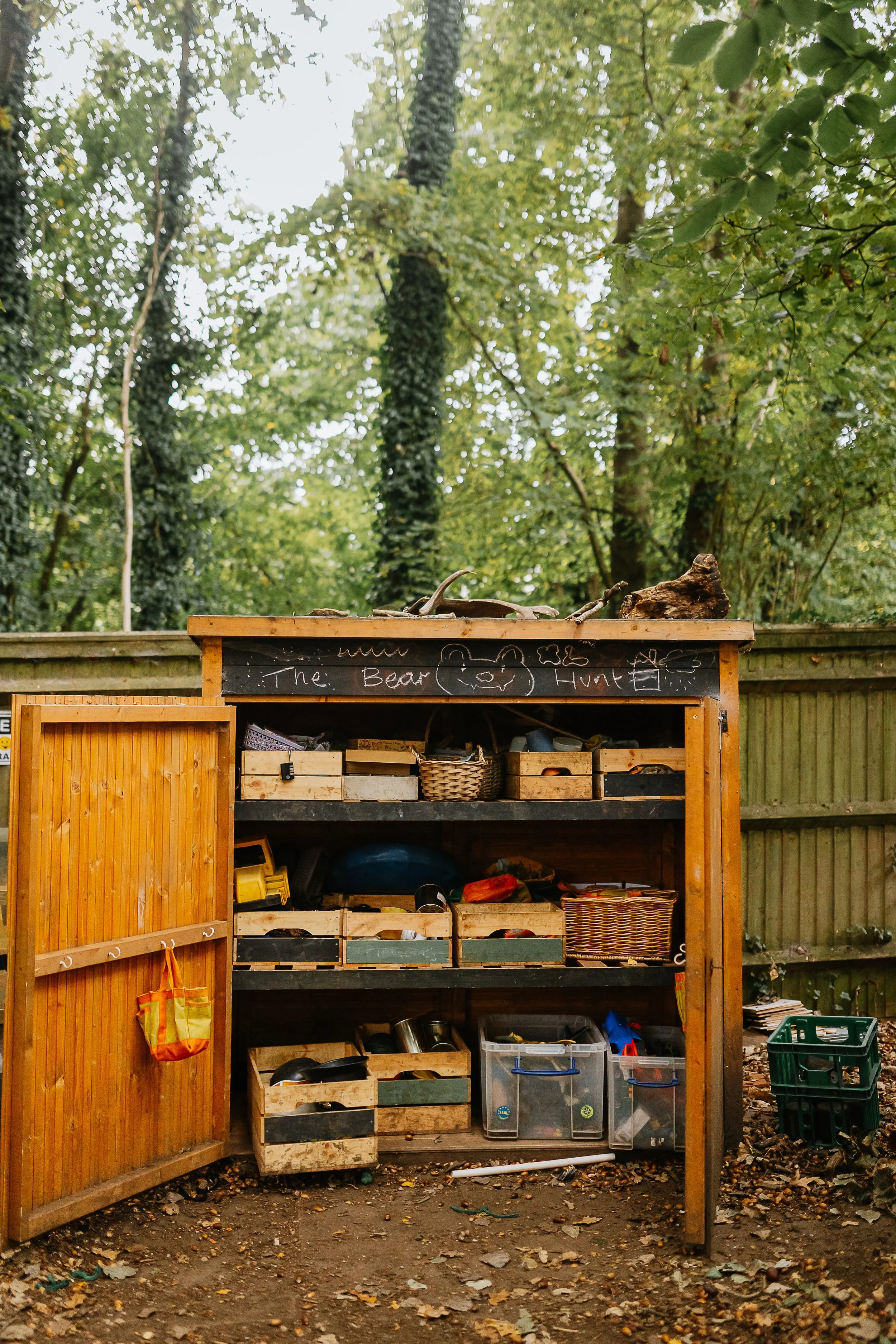 An outdoor wooden shed with open doors revealing storage bins and garden tools inside. The shed is situated in a backyard with a wooden fence, trees, and fallen leaves on the ground.