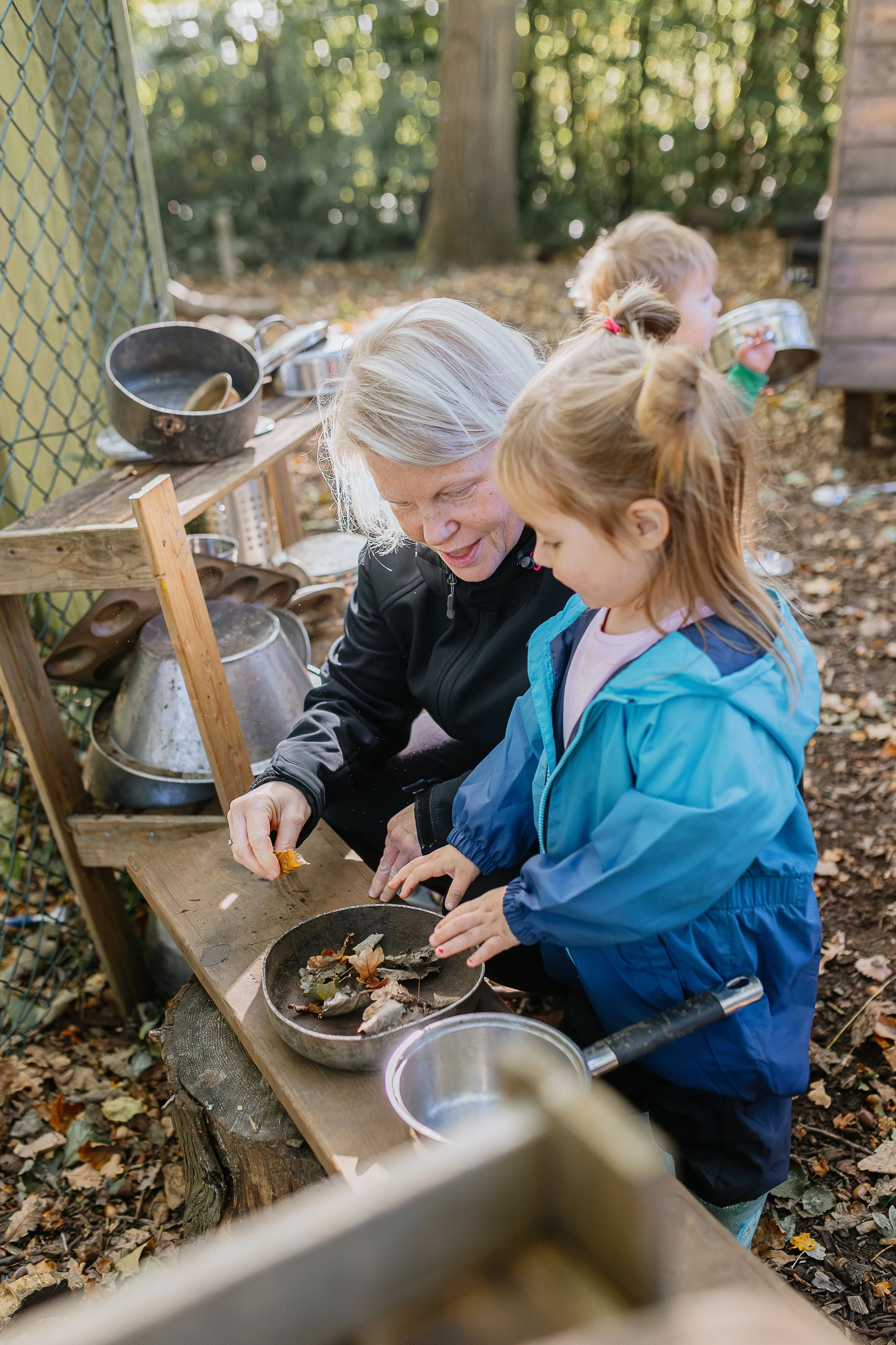 An outdoor scene in a wooded area with a woman and three children collecting leaves and natural items in a black tray, with additional pots and utensils on a wooden shelf.
