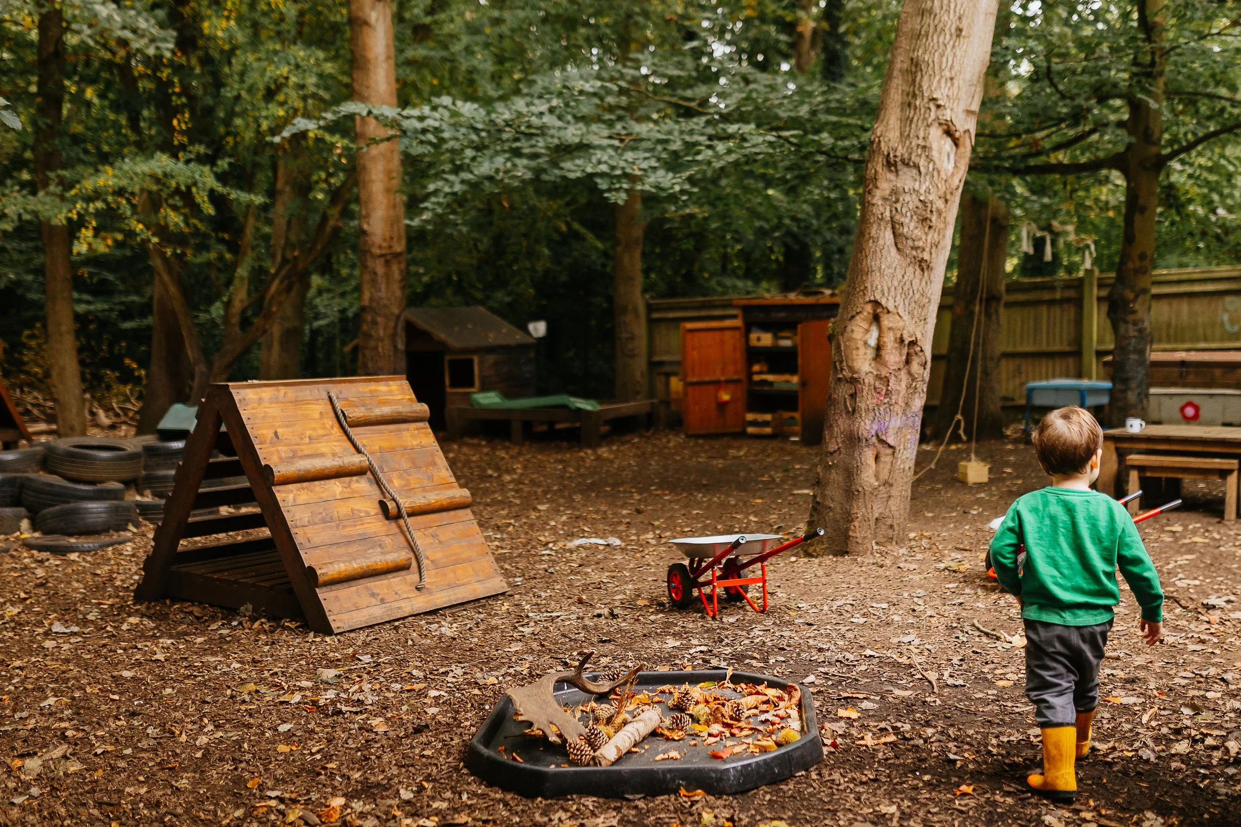 A backyard with trees, wooden play structures, a small shed, and a child in a green hoodie and yellow rain boots walking away from the camera. There are tires stacked on the left and some toys and tools around.