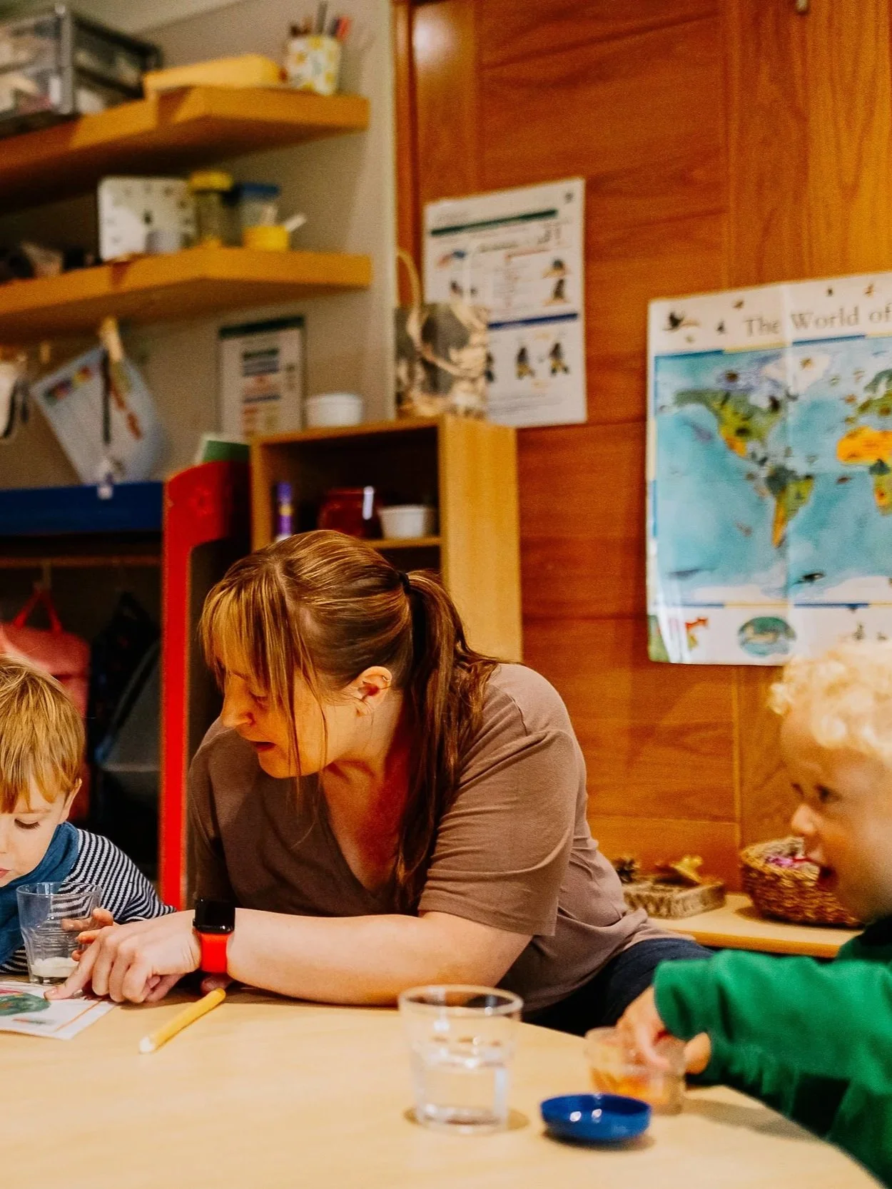 A woman with brown hair tied in a ponytail is sitting at a table with two young children, one with blonde hair and the other with red hair. They are engaged in an activity involving small glasses and containers, possibly a science experiment or craft. The background includes wooden wall paneling, world maps, posters, and shelves with supplies.