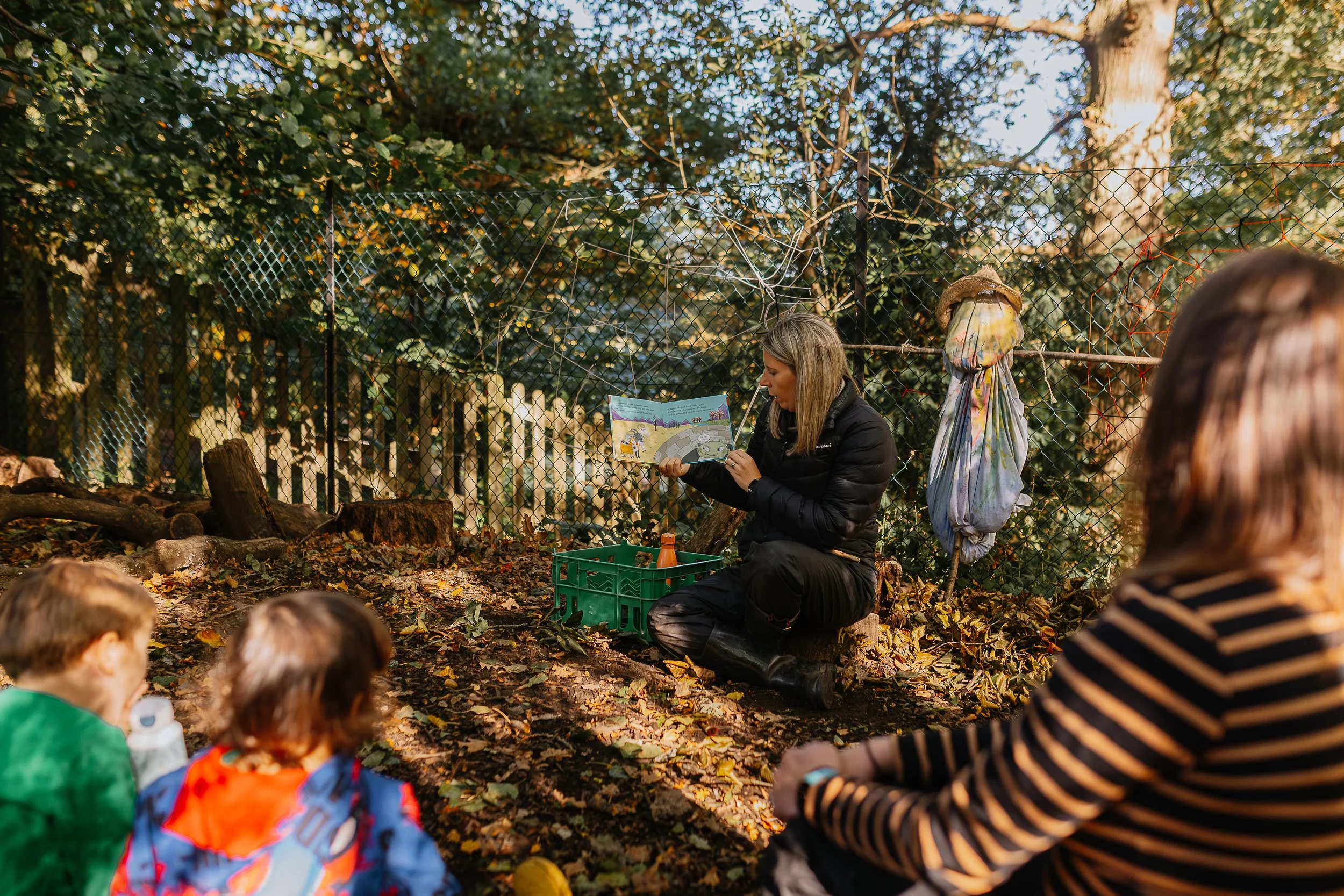 A woman reading a book to children outdoors in a wooded area with fallen leaves, behind a chain-link fence with trees and sunlight in the background.