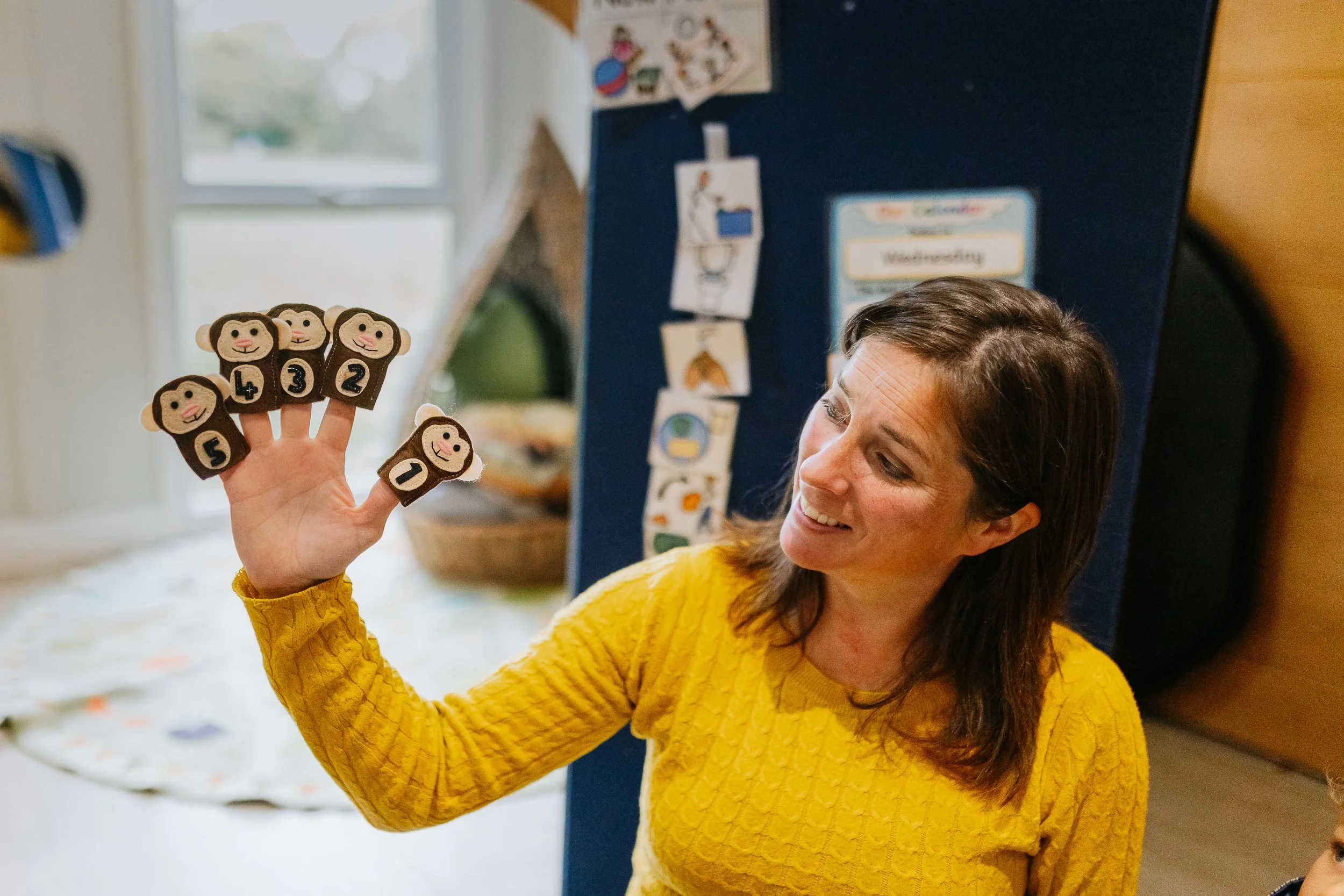 A woman in a yellow sweater holding up five animal-shaped hand puppets with numbers on them, smiling and looking at the puppets.