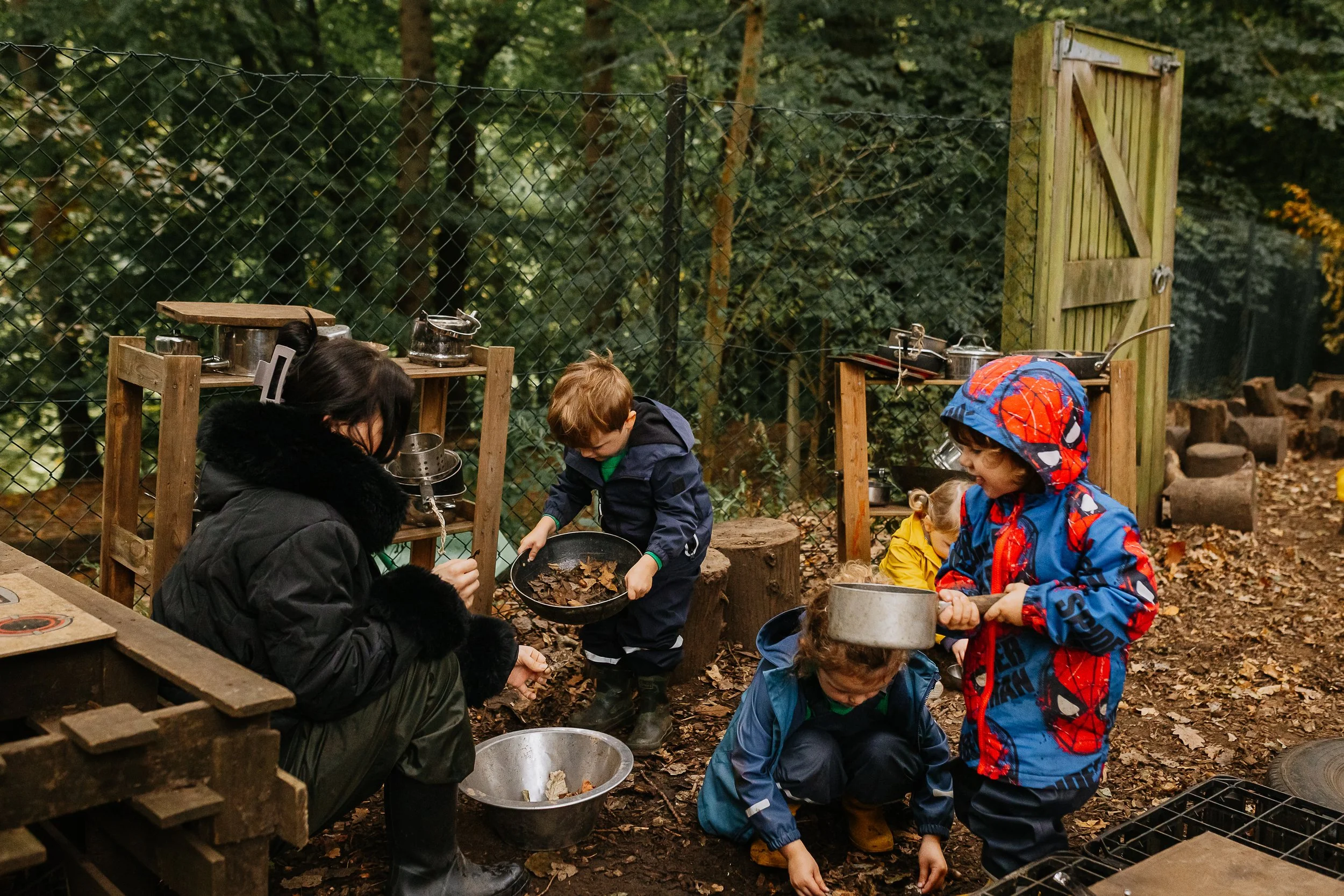 Children and an adult outdoors in a wooded area, gathered around cooking equipment, preparing food with leaves scattered on the ground, behind a chain-link fence and trees.