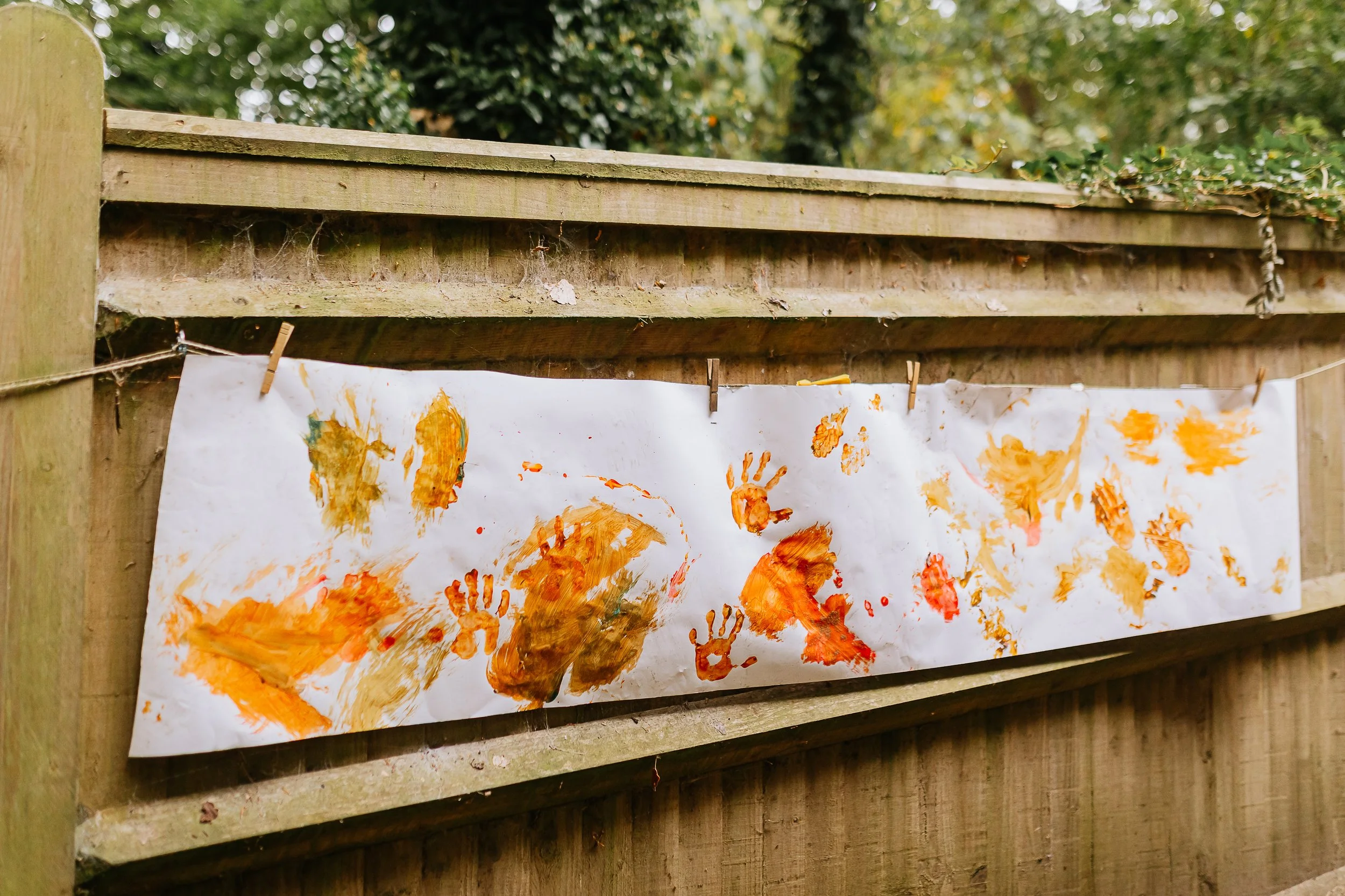 A piece of outdoor string art with painted handprints and handprints painted in orange and yellow on white paper, hanging on a dog-eared wooden fence in a backyard with trees in the background.