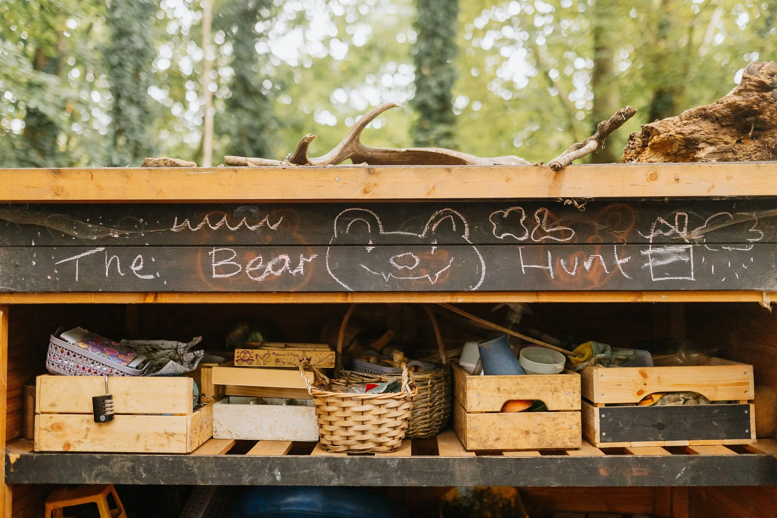 Wooden shelf with various small baskets and containers, surrounded by logs and branches, with a sign displaying a pumpkin, clouds, and the words "The Beanstalk" and "Hunting" underneath, set outdoors among trees.