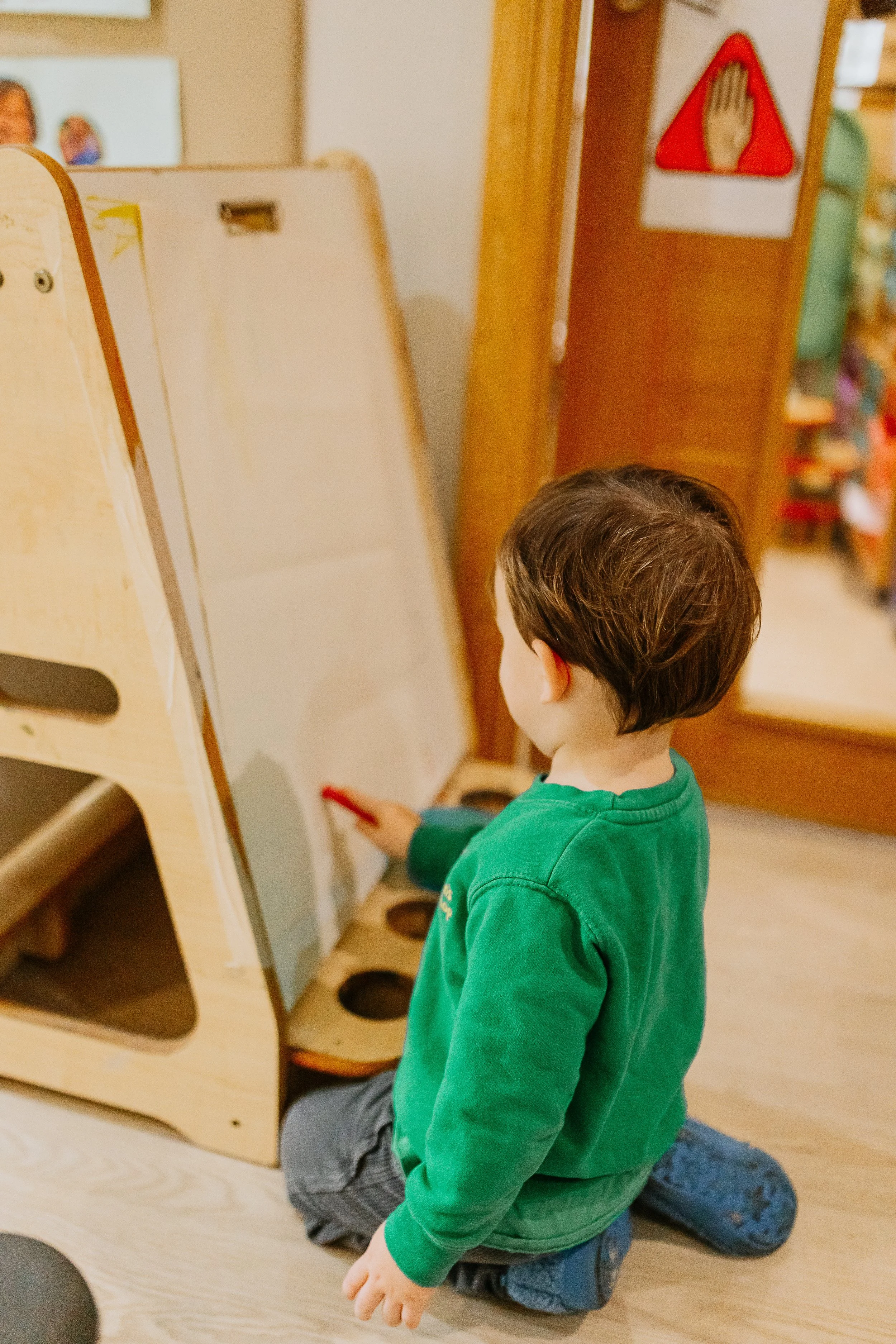 A young boy with brown hair, wearing a green hoodie, kneeling on the floor in a daycare or preschool setting. He is holding a red marker and working on a whiteboard or drawing surface. There are circular holes in a wooden panel in front of him and a red and white warning sign on the wall behind.
