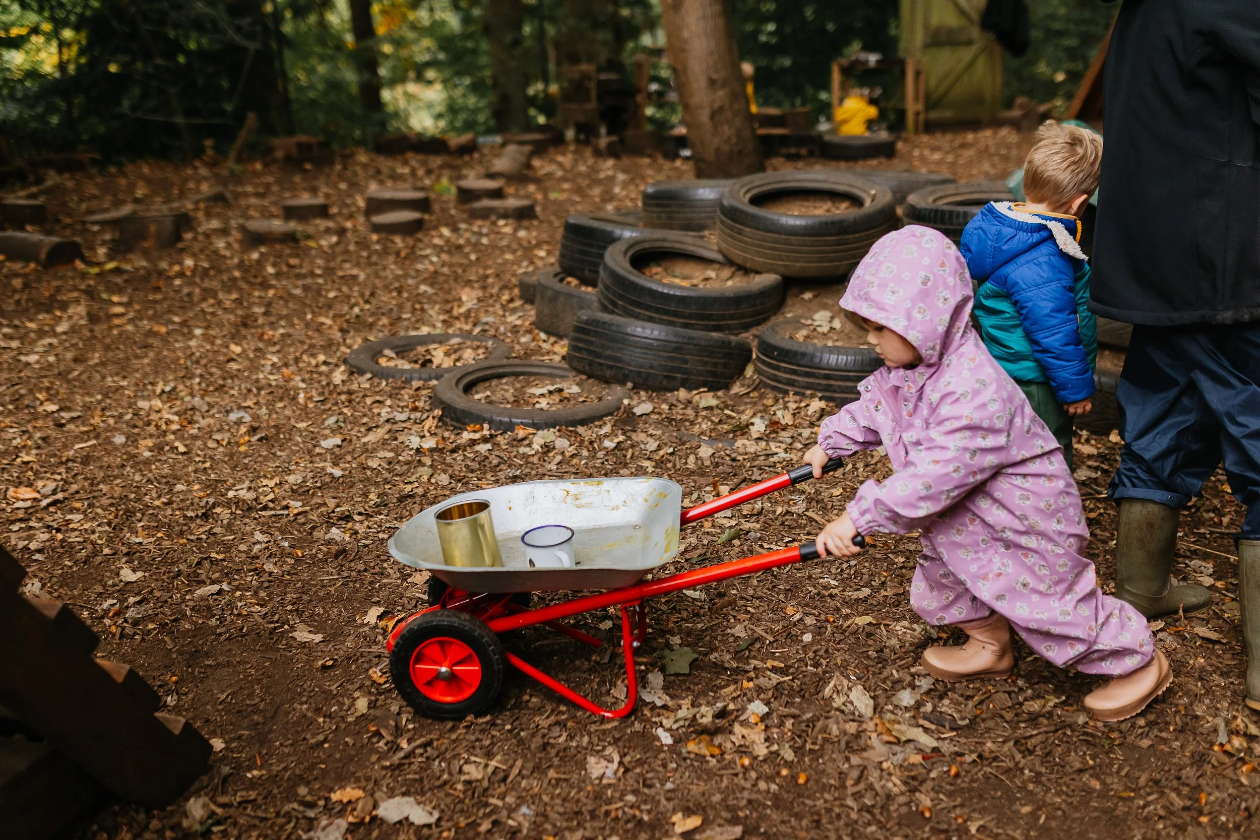 Two young children, a girl in a pink hooded jacket and a boy in a blue jacket, playing outdoors near stacked tires and a red toy wheelbarrow filled with metal cups.
