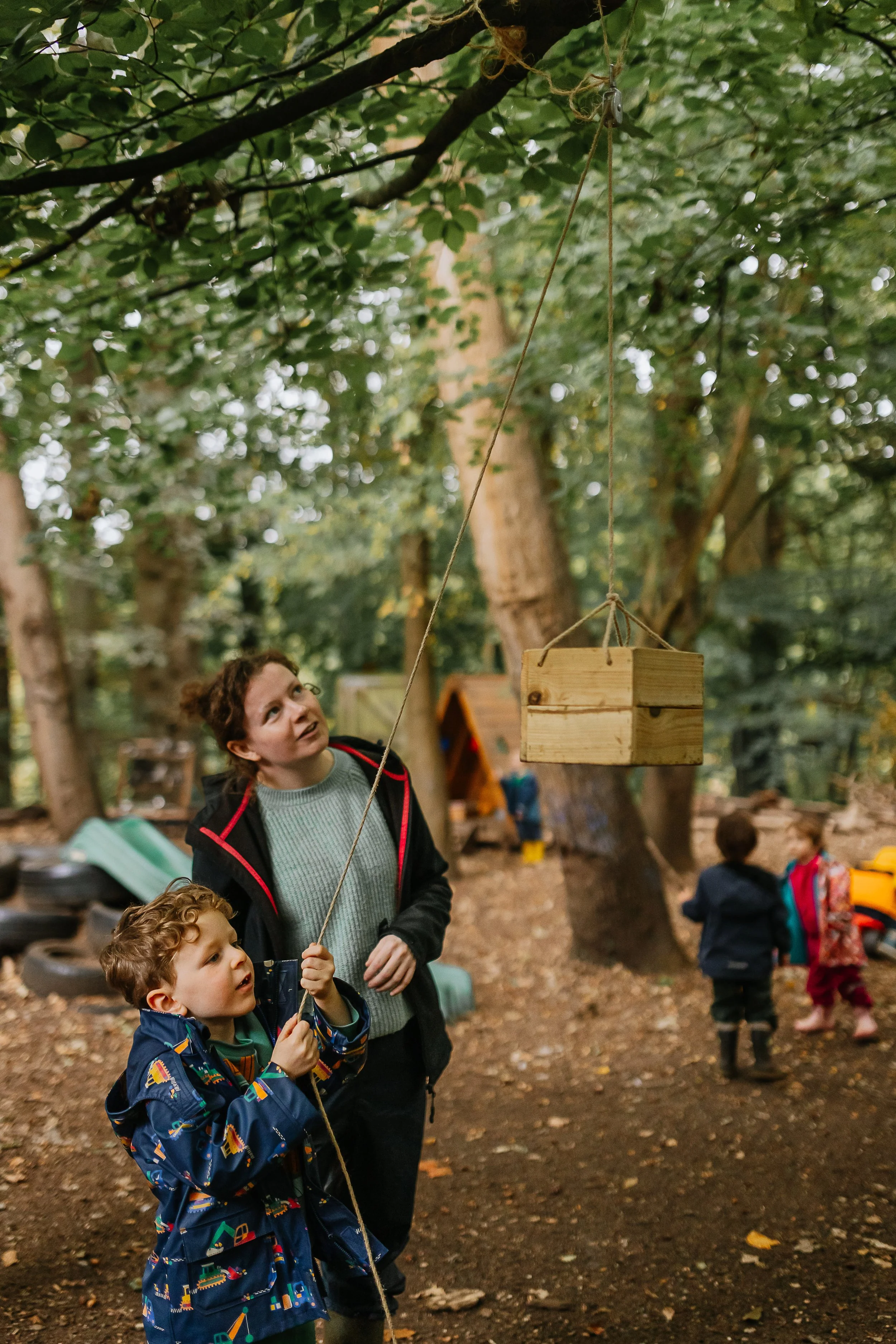 A woman and children in a wooded outdoor area, with a small wooden hanging box suspended by a string, and trees in the background.