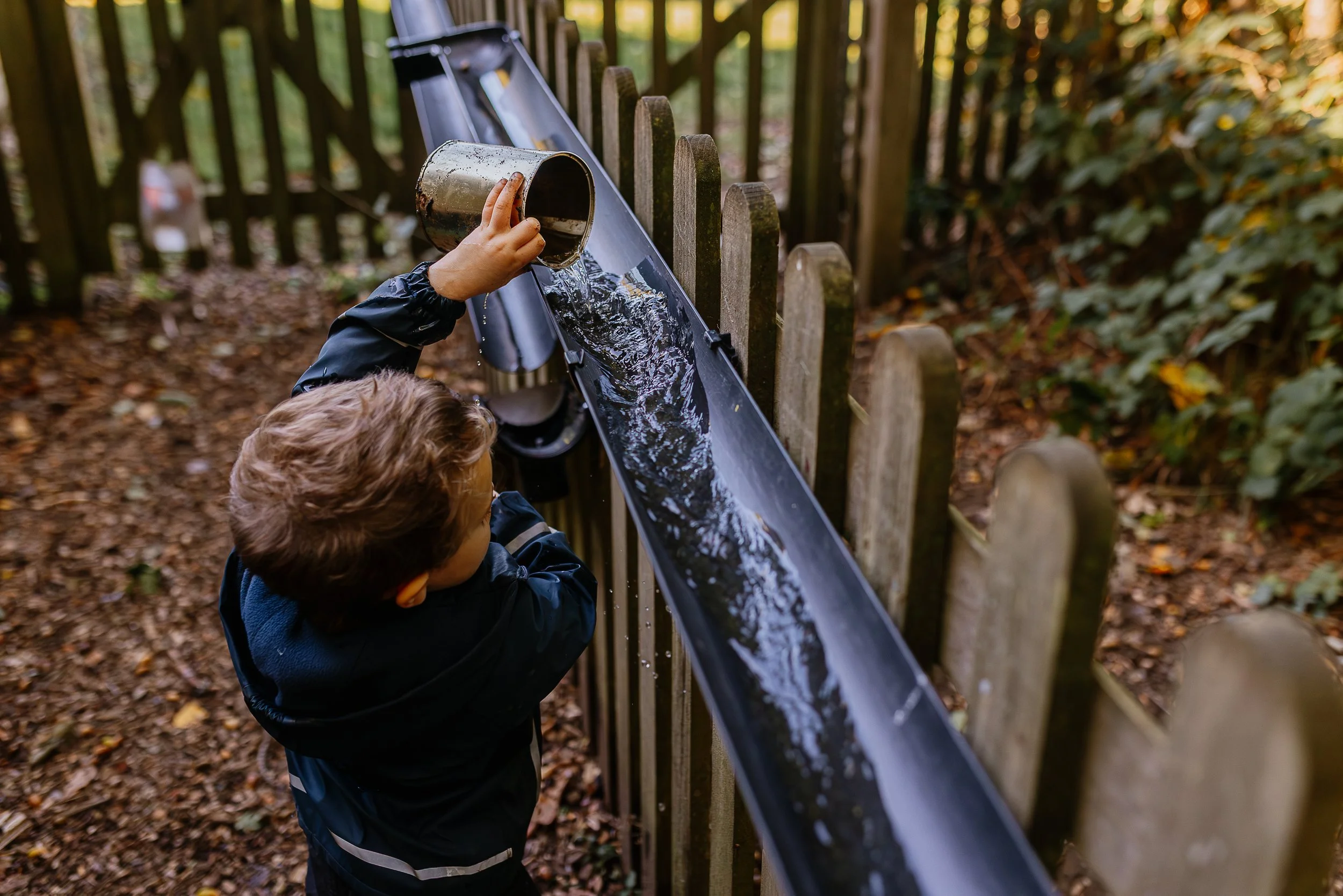 Two children are playing at a black metal slide in an outdoor park. One child is holding a metal container, pouring water onto the slide, while the other child looks on. The setting features a wooded area with trees and fallen leaves.