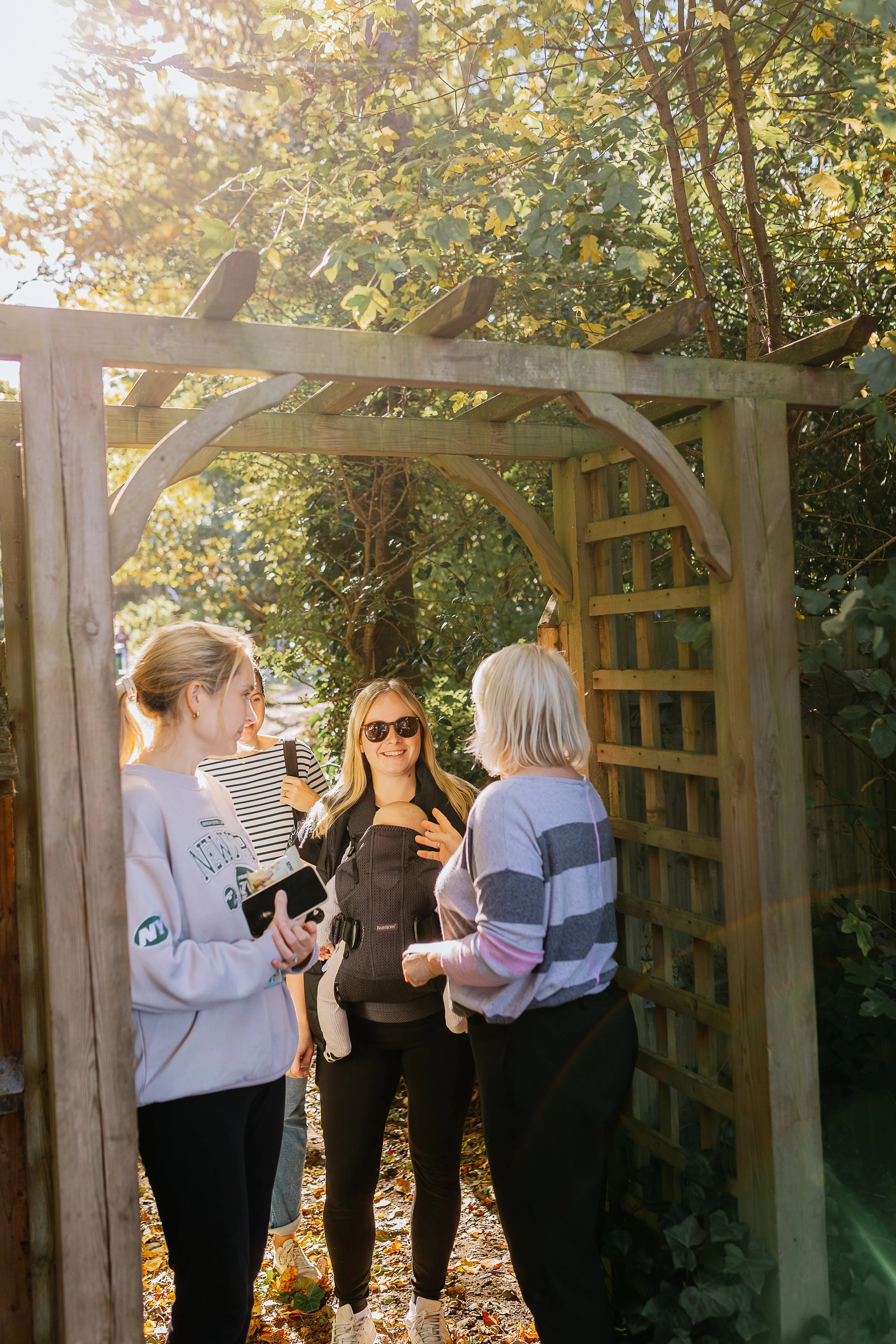 Group of women standing under a wooden archway outdoors, engaged in conversation, in a garden or park area with trees and sunlight.