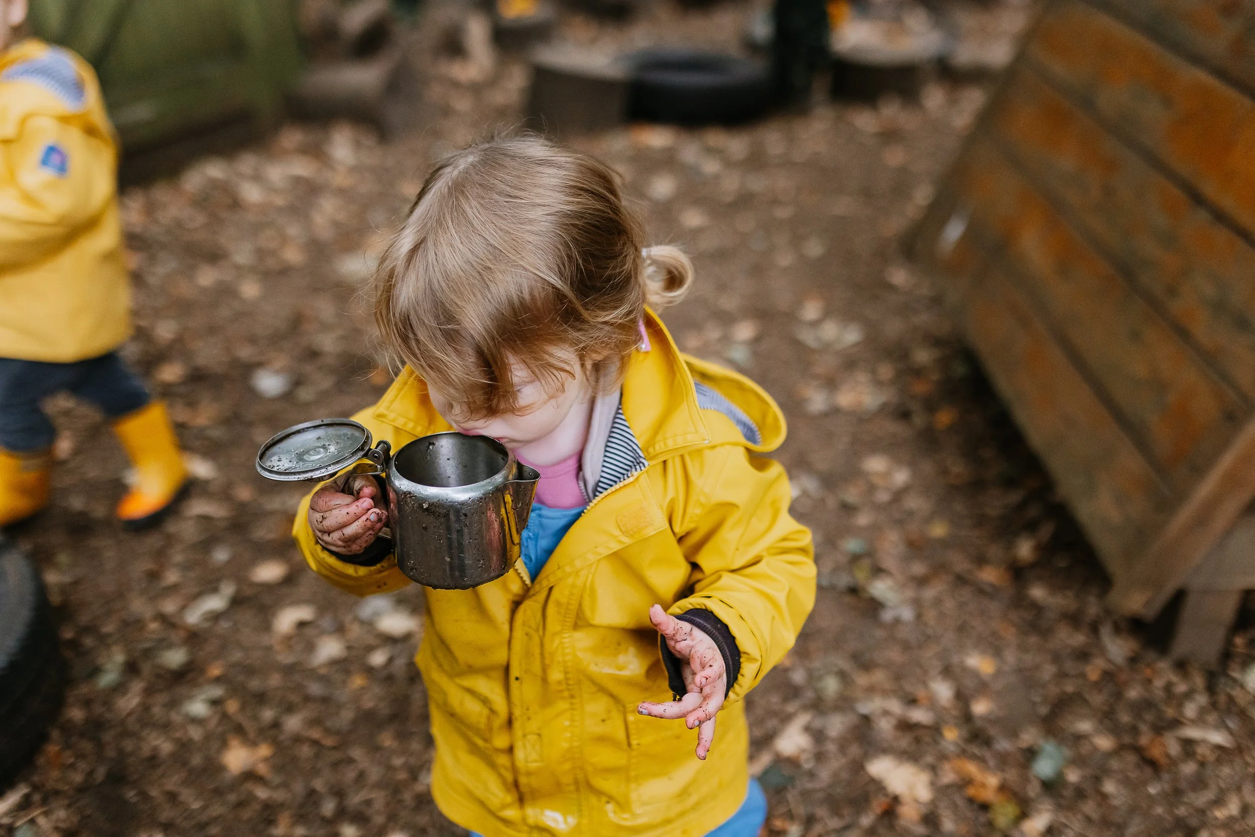 Young child with curly blonde hair wearing a yellow raincoat holding a metal cup, standing outdoors on a leaf-covered ground.