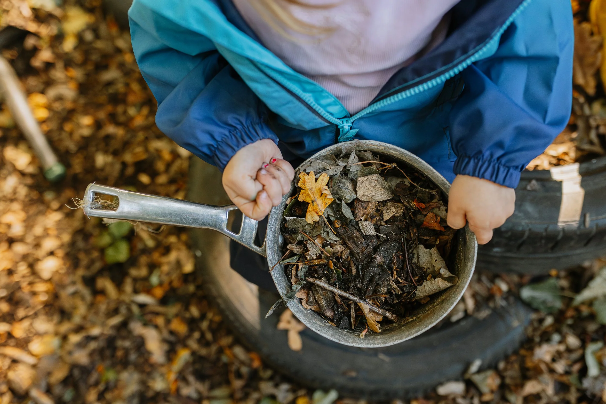 A child wearing a blue jacket holding a metal shovel filled with fallen autumn leaves, dirt, and small sticks, surrounded by a ground covered with more leaves.