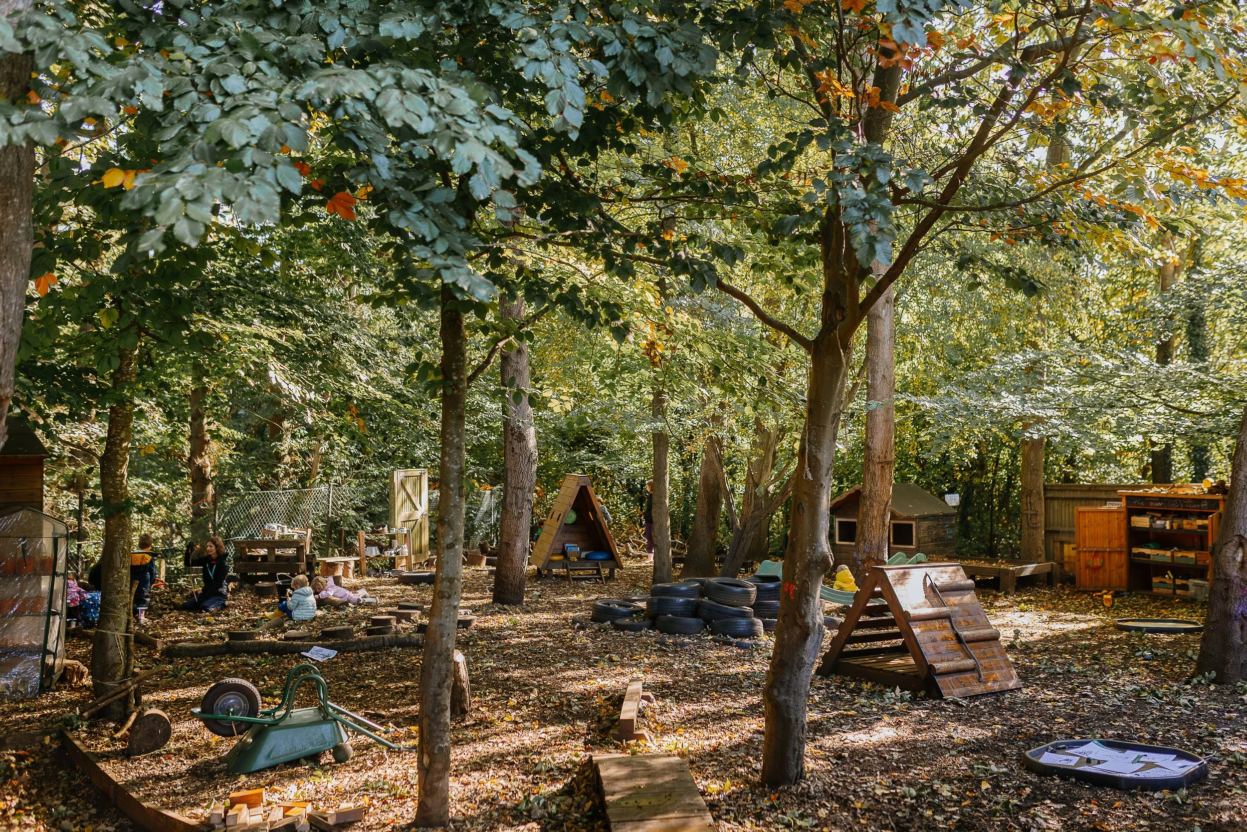 A backyard playground area with tall trees, children playing, and various wooden play structures and outdoor toys scattered on the ground.
