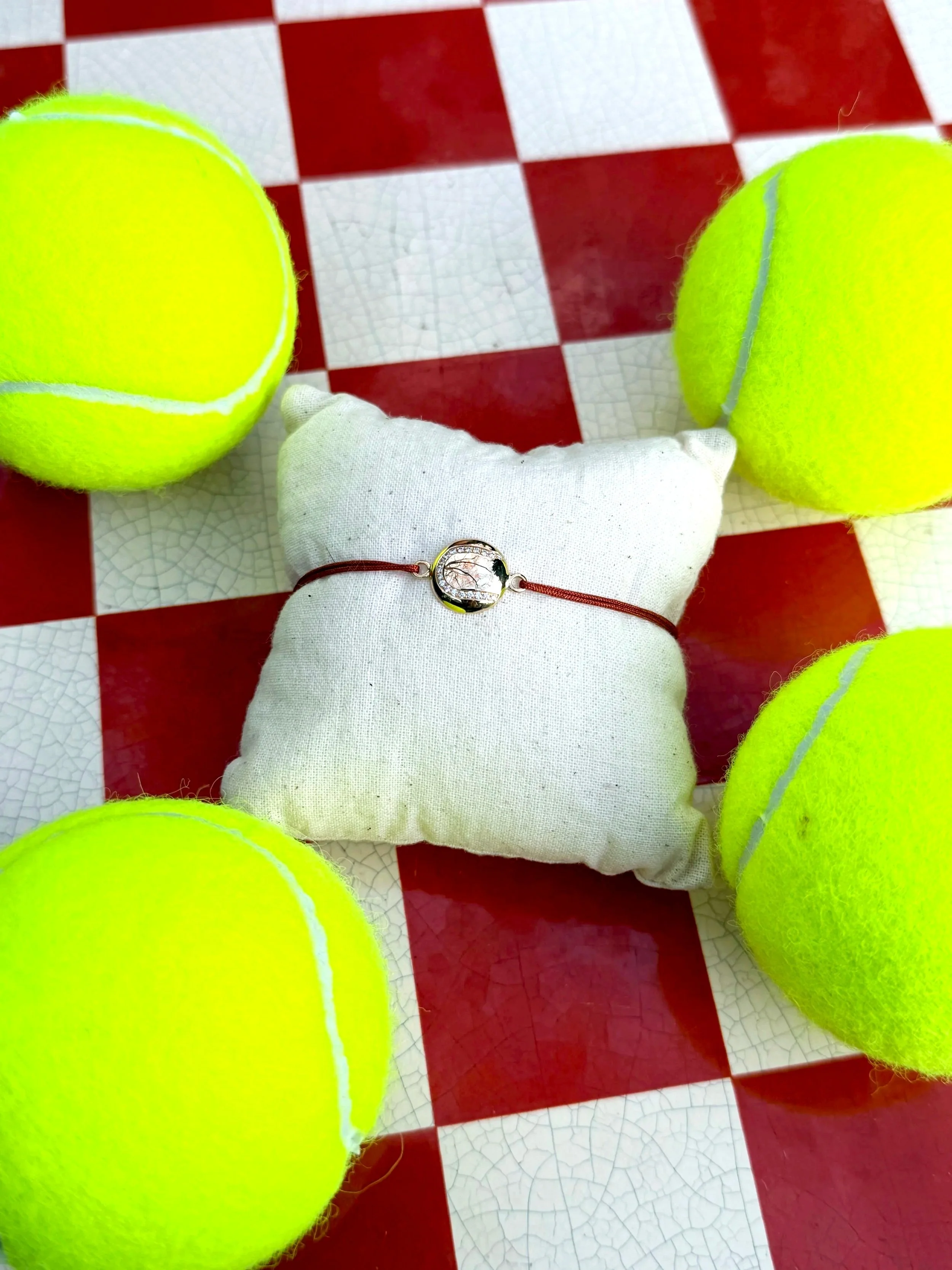 Bracelet balle de tennis en or et diamants  placées sur un duvet blanc, à côté de plusieurs autres balles de tennis, sur un sol en mosaïque rouge et blanche.
Joaillerie, bijoux , diamants, or 18 carats . 
Roland Garros, Wimbledon