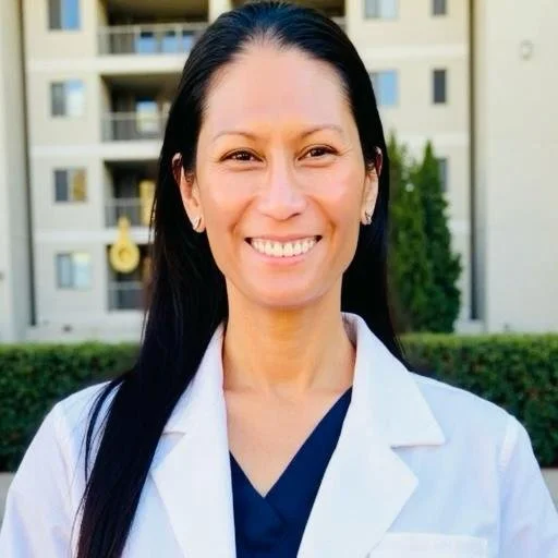 A woman with long dark hair smiling outdoors with apartment buildings in the background, wearing a white coat and navy blue scrubs.