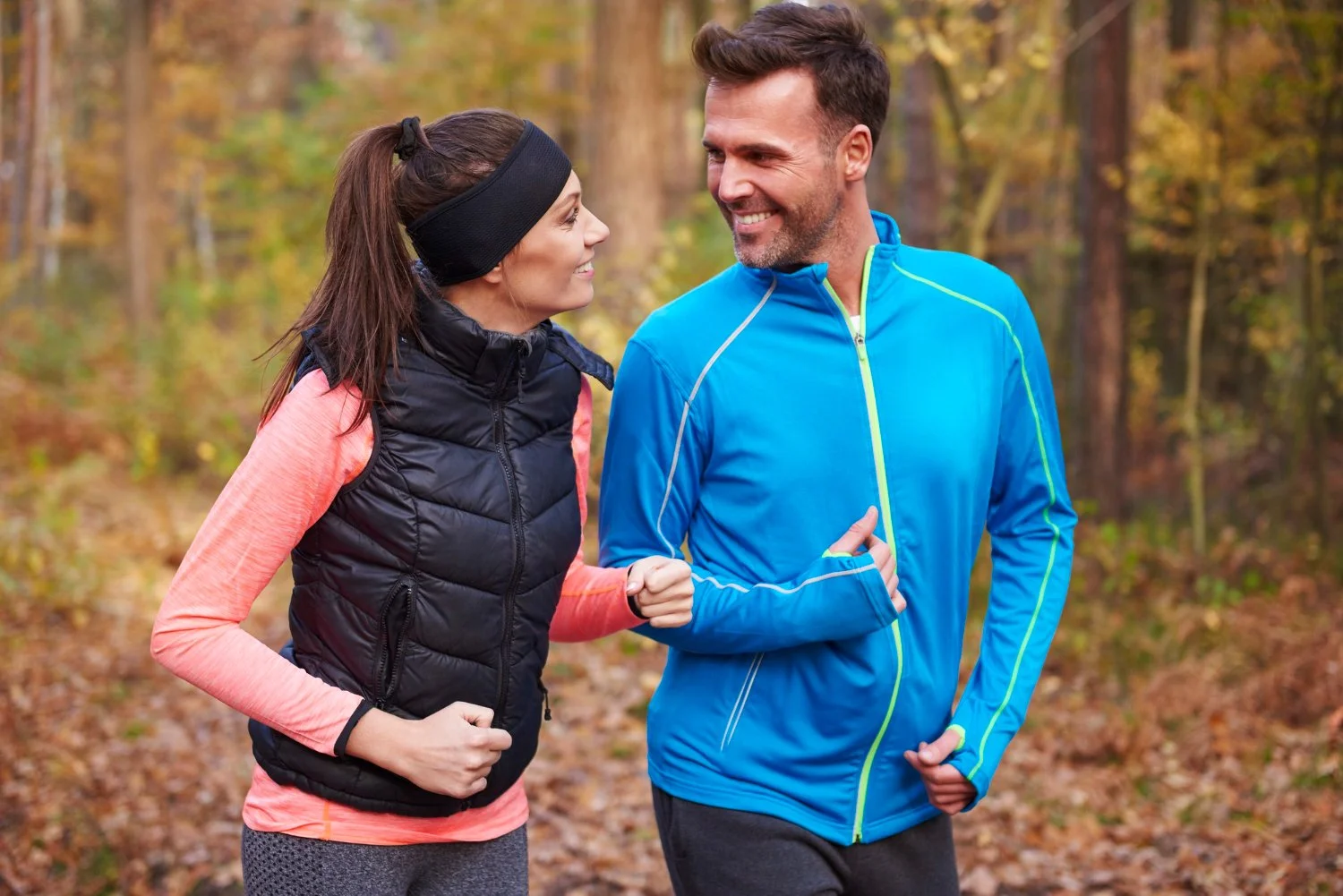 A smiling man and woman jog together in a forest during autumn, wearing athletic gear.