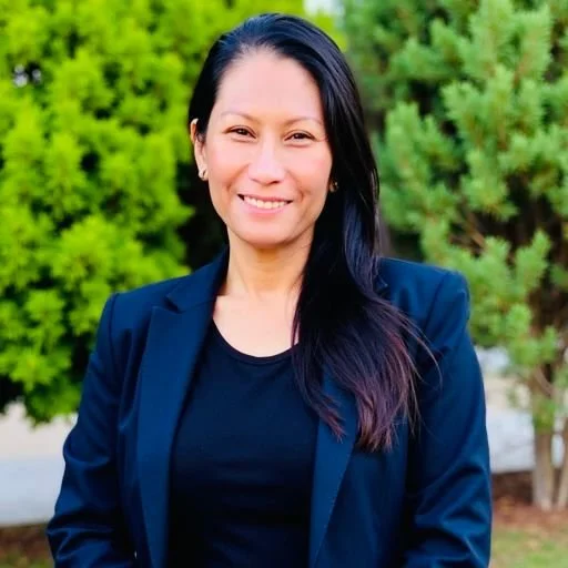 A woman with long dark hair smiling in front of green trees, wearing a navy blazer and black shirt.