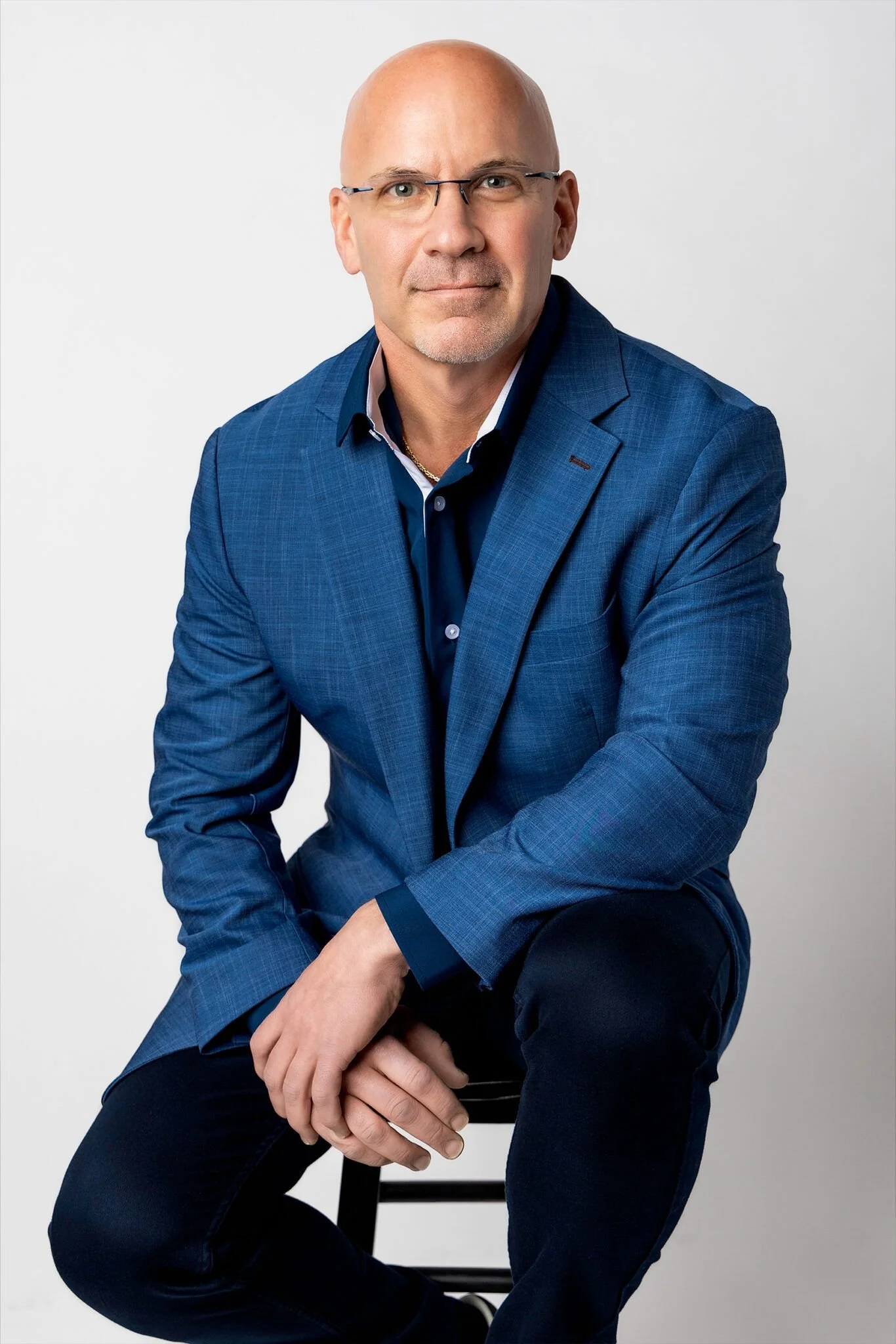 A middle-aged man wearing glasses, a blue checked blazer, and a dark blue shirt, sitting on a chair against a plain white background.