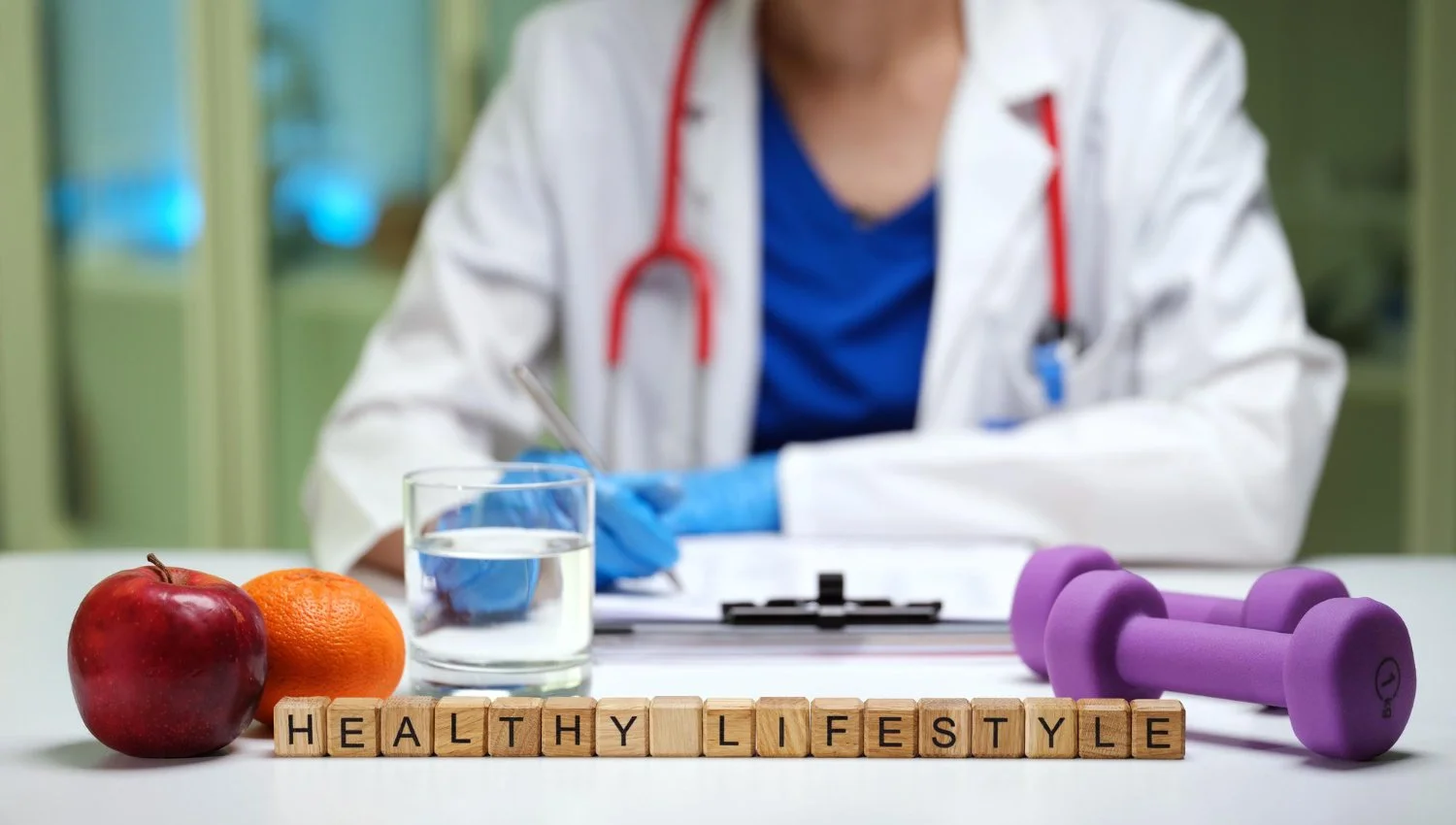 A doctor or healthcare professional sitting at a table with health-related items including a red apple, an orange, a glass of water, purple dumbbells, and wooden blocks spelling 'HEALTHY LIFESTYLE'; the person is wearing a white coat, blue scrubs, and a stethoscope around their neck.