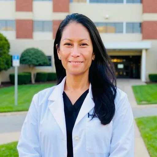 A woman in a white medical coat standing outside in front of a building with green bushes and grass.