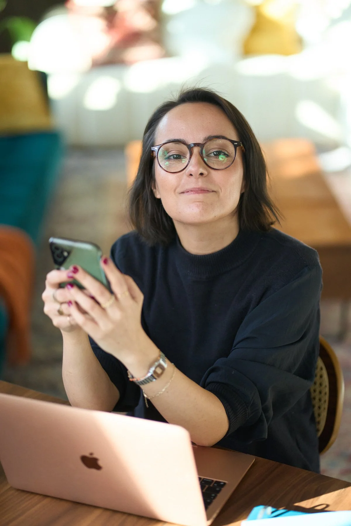 A woman with glasses sitting at a table with a pink MacBook and holding a smartphone, in a brightly lit cafe or workspace.