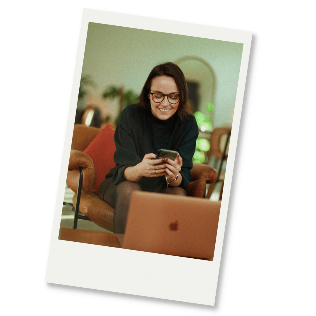 Une femme souriante regarde son téléphone portable dans un salon confortable, avec un ordinateur portable sur la table devant elle.