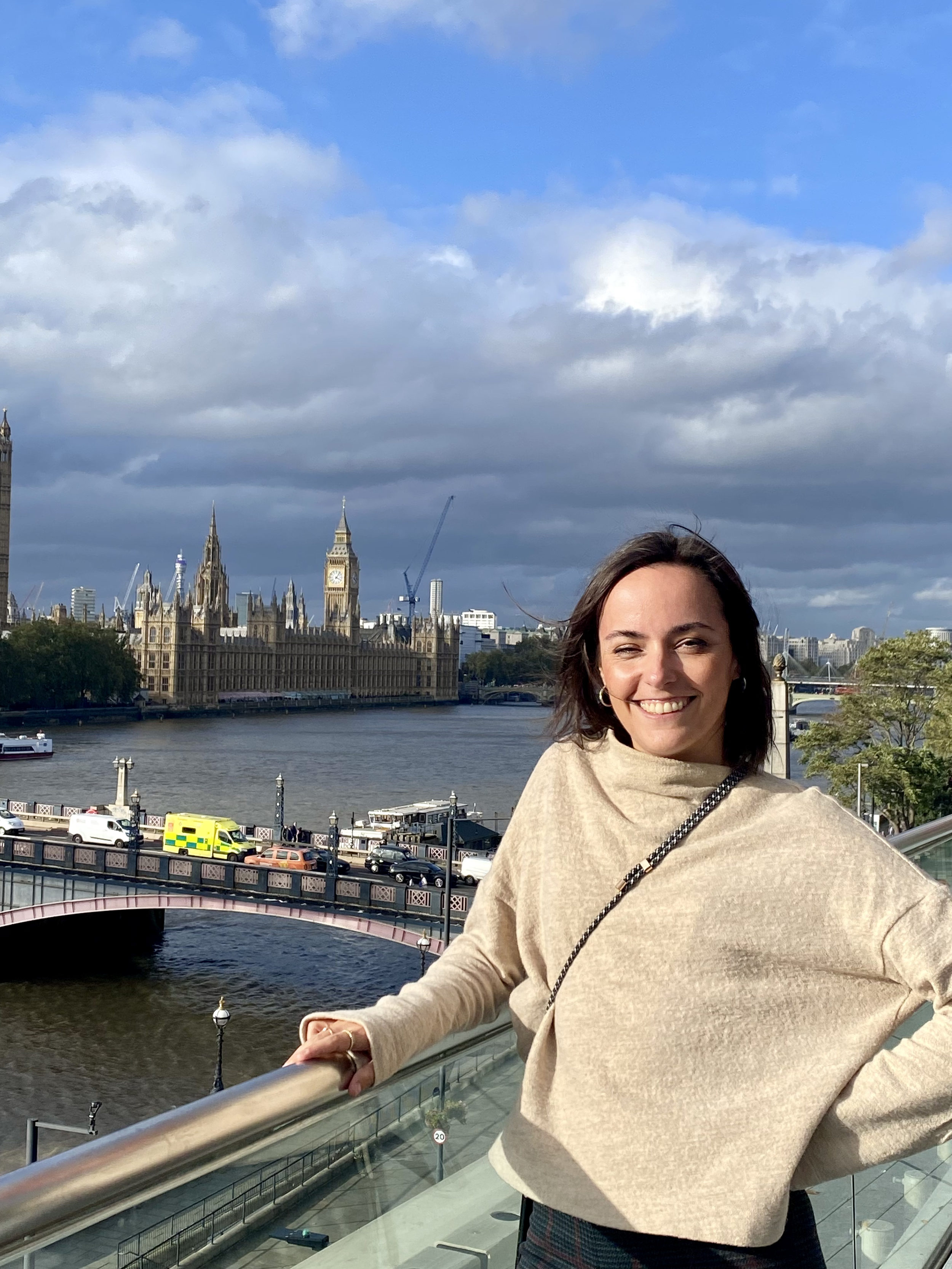 Une femme souriante portant un manteau beige se tient près de la rivière Thames à Londres avec le Parlement et Big Ben en arrière-plan.