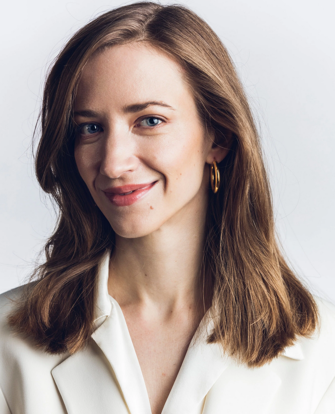 Porträt einer lachenden Frau mit braunen, geschwungenen Haaren und blauen Augen, tragen goldene Creolen, vor hellem Hintergrund.