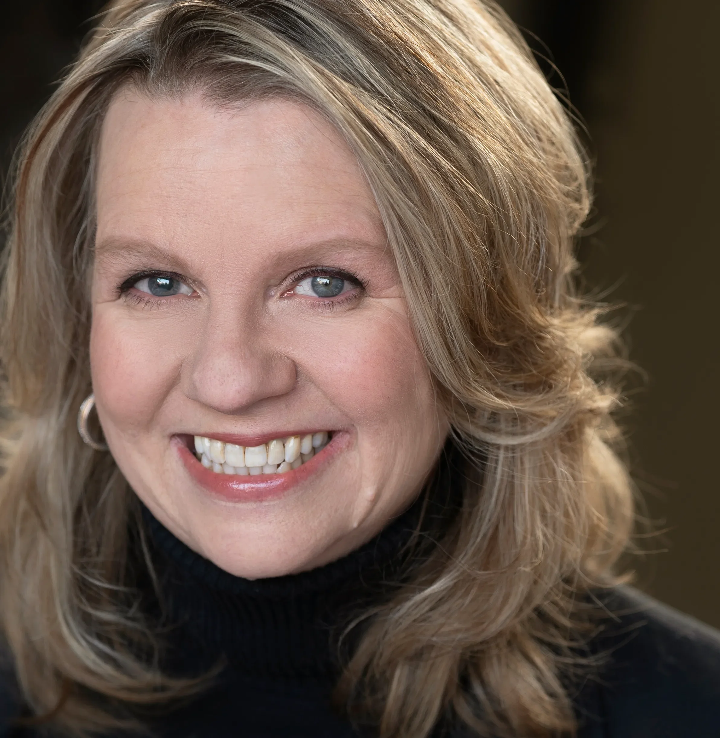 Nahaufnahme eines lächelnden schwarzen Frauen mit blauen Augen, mittellangen, gewellten, blond-braunen Haaren, trägt einen schwarzen Rollkragenpullover und Silberschmuck.