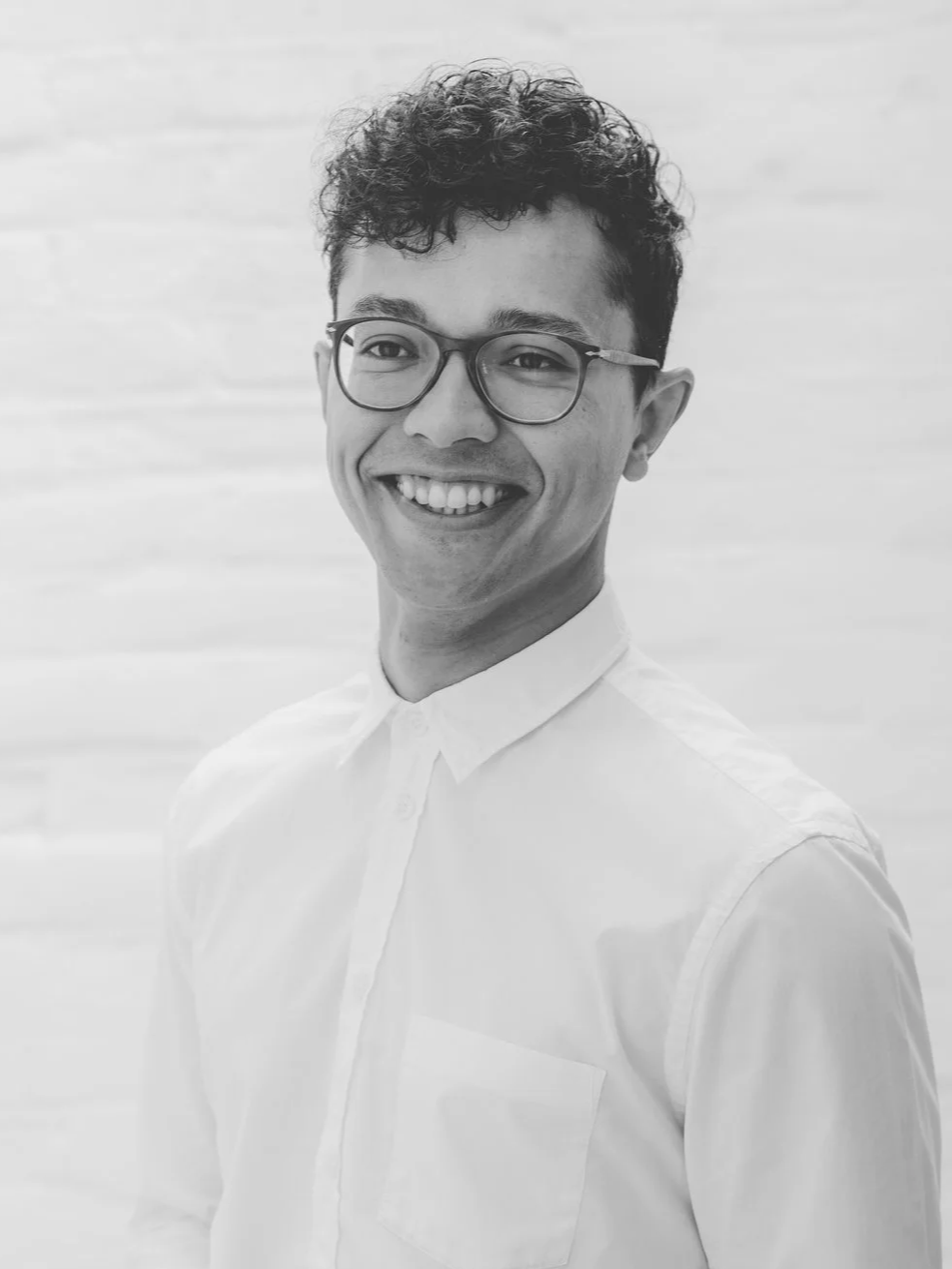Black and white portrait of a smiling man wearing glasses and a white button-up shirt, standing against a light-colored textured background.