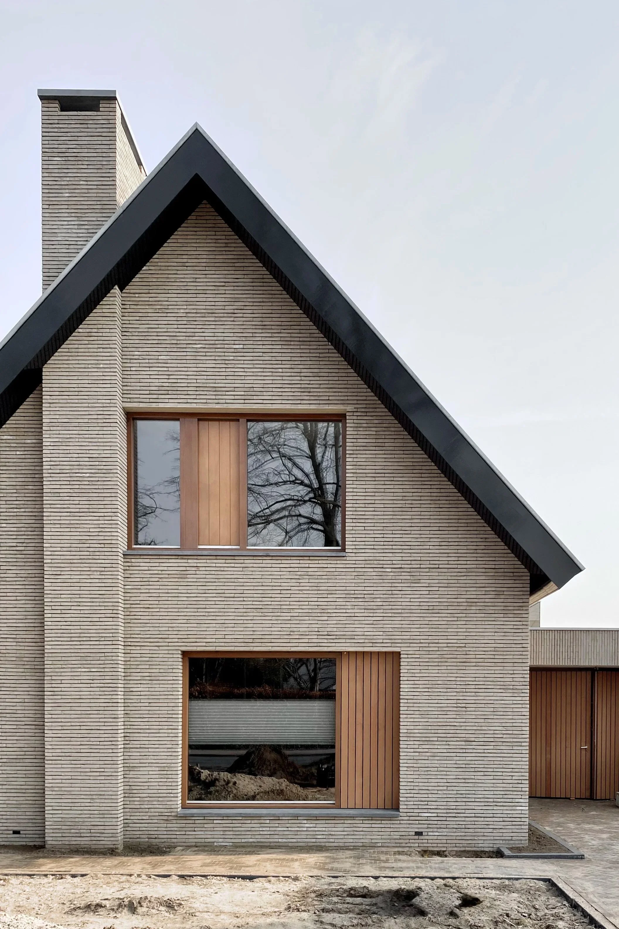 Modern house with beige brick exterior, large rectangular window with wooden framing, sloped roof, and chimney, under cloudy sky.