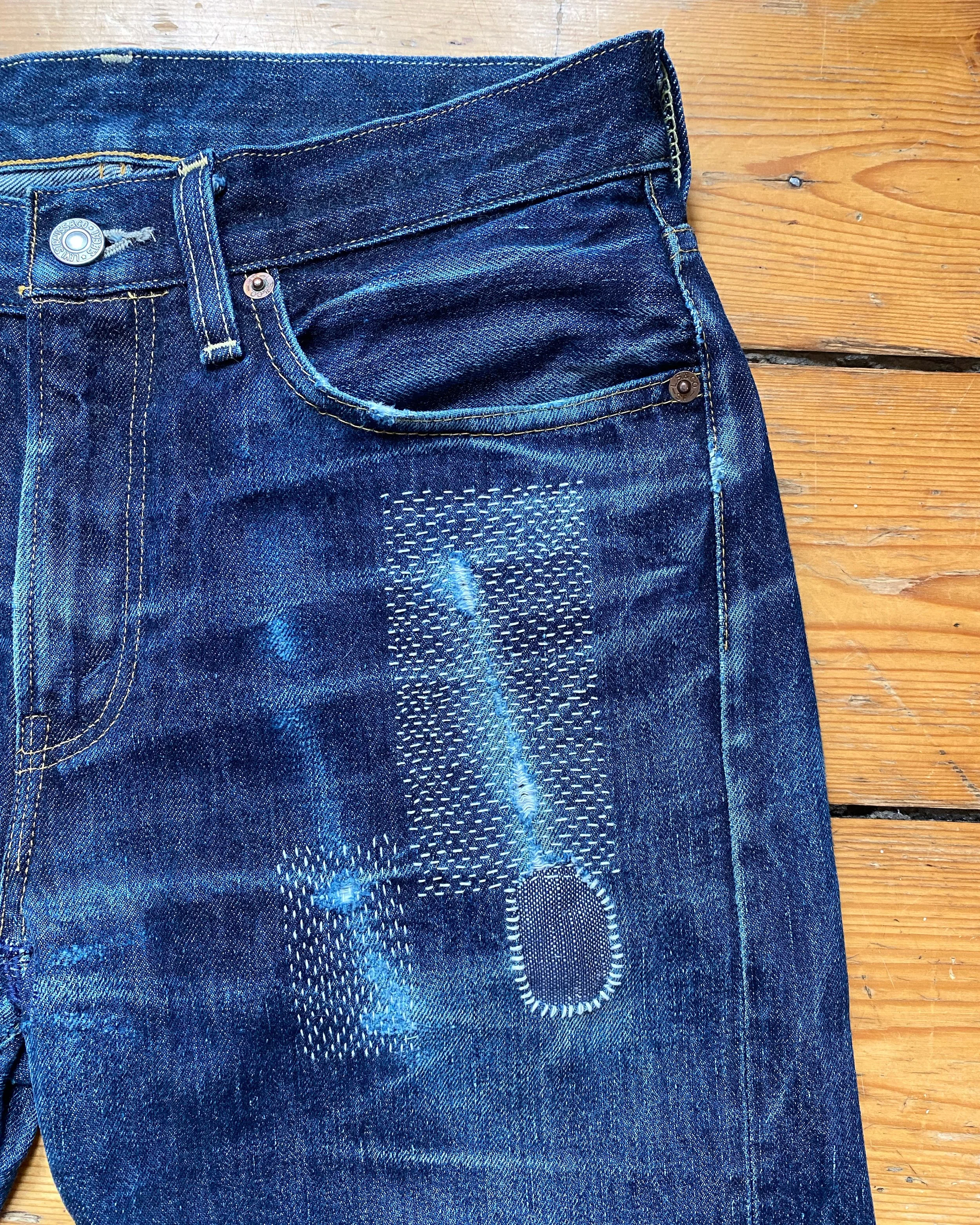 Visible mend on denim: Jeans have been patched on reverse near front pocket and reinforced with visible sashiko stitching and whip stitching around the edge of a hole, all in white embroidery thread.
