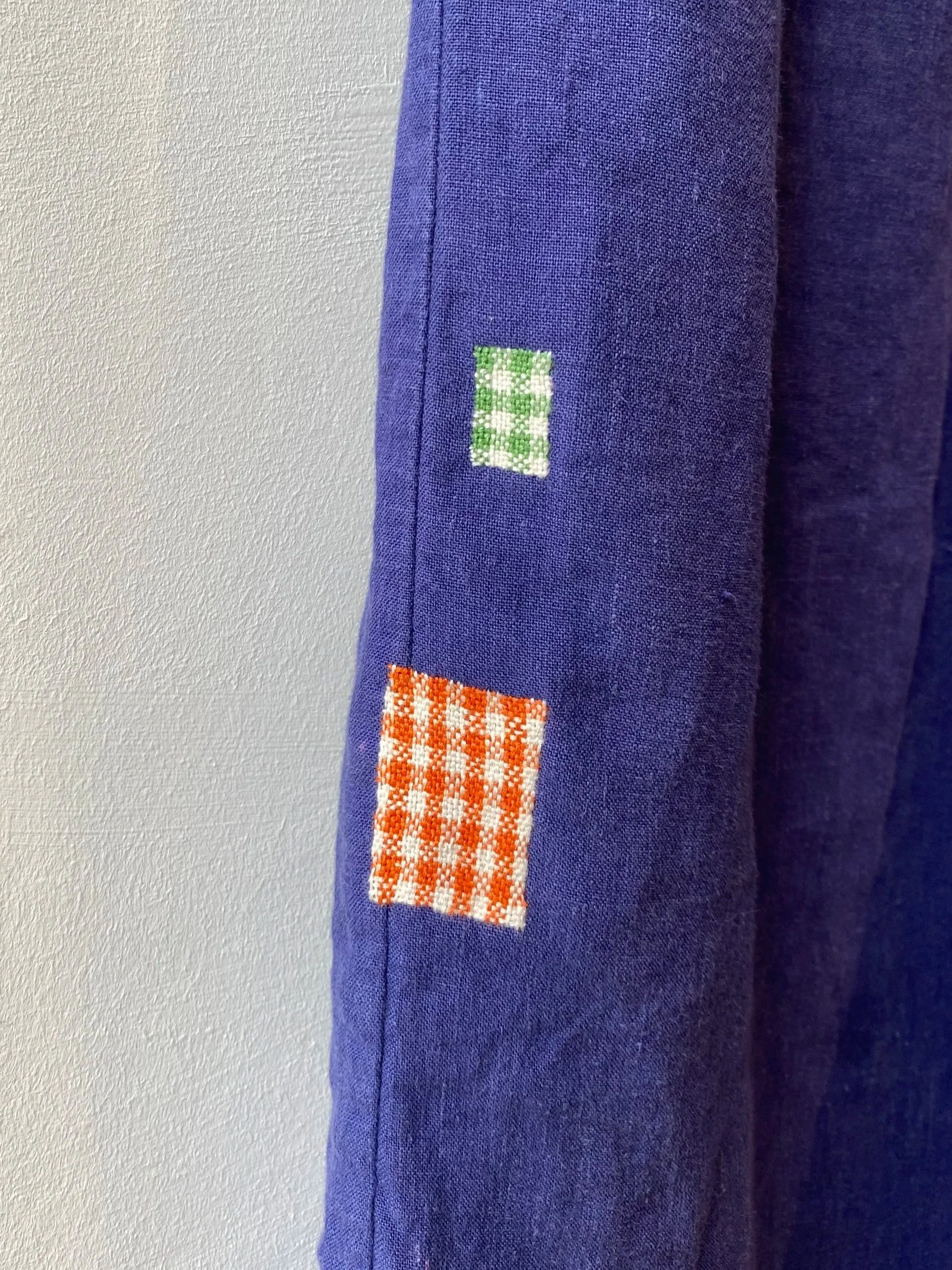 Visible darn on linen trousers: Violet trousers with two different size square darns in a gingham pattern, one in orange and cream, the other in apple green and cream