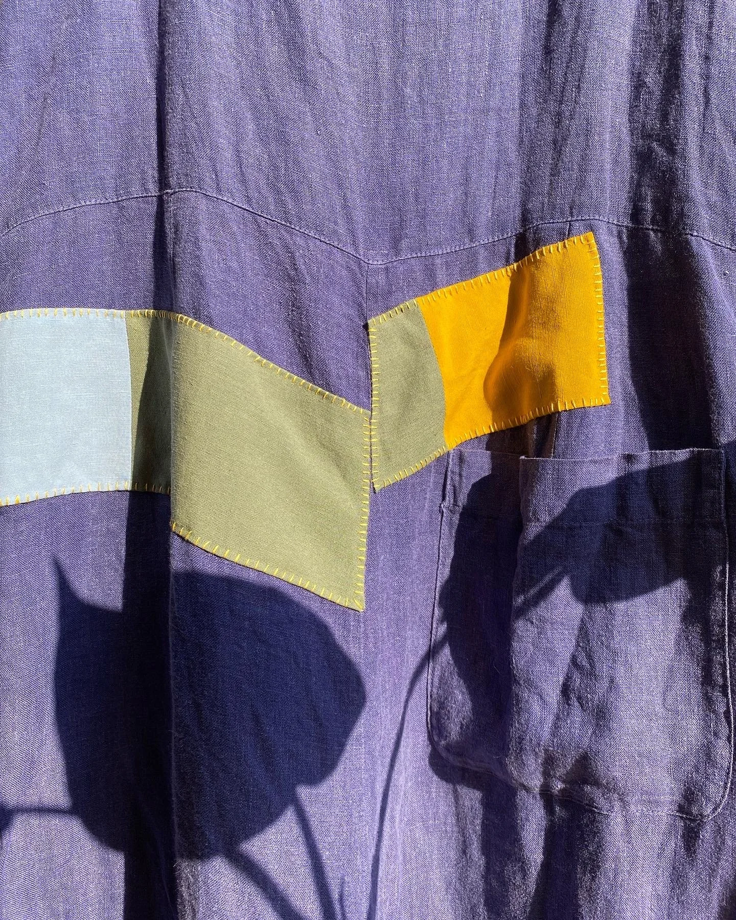 Visible patched mend on linen: Yellow, sage green and light blue patchwork shapes stitched onto soft purple linen in a yellow thread