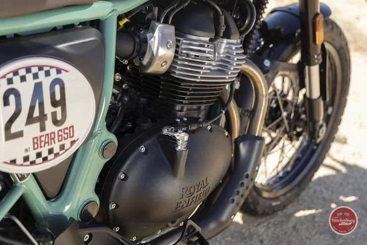 Close-up of a vintage Royal Enfield motorcycle with a green frame, showing its engine, part of the wheel, and a racing number badge reading 249 with 'inter' and 'BEAR 650'.