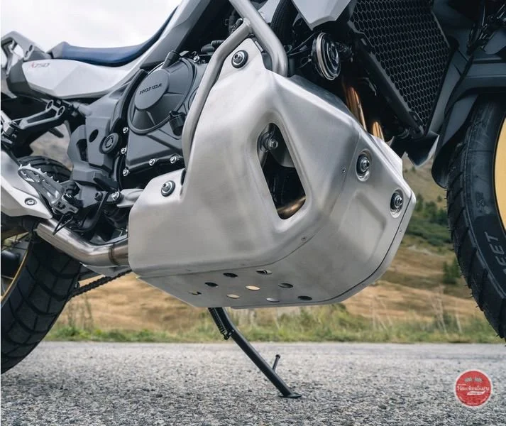 Close-up of a motorcycle engine and exhaust system, showing a large metallic exhaust cover, with a paved road and grassy area in the background.