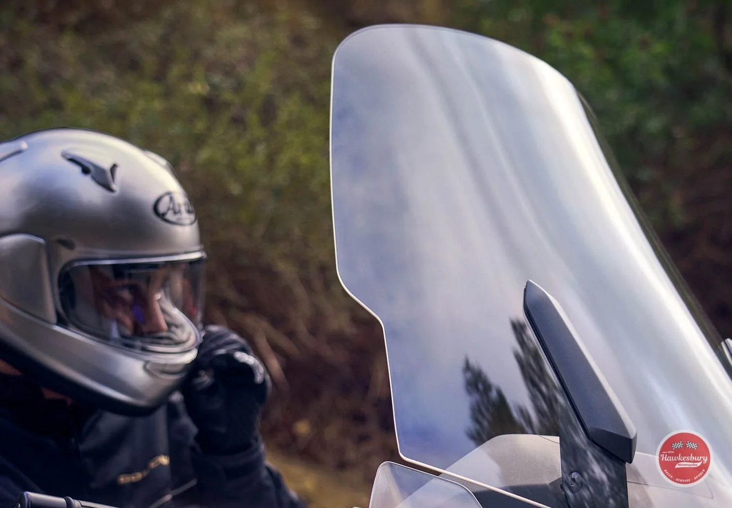A motorcyclist wearing a silver helmet and black riding gear, sitting on a motorcycle next to a windscreen with a blurred natural background.