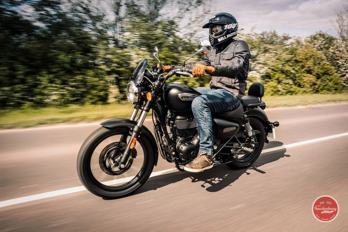 A person riding a black Royal Enfield motorcycle on a road with blurred trees in the background. The rider is wearing a black helmet, gray jacket, brown gloves, blue jeans, and brown shoes.