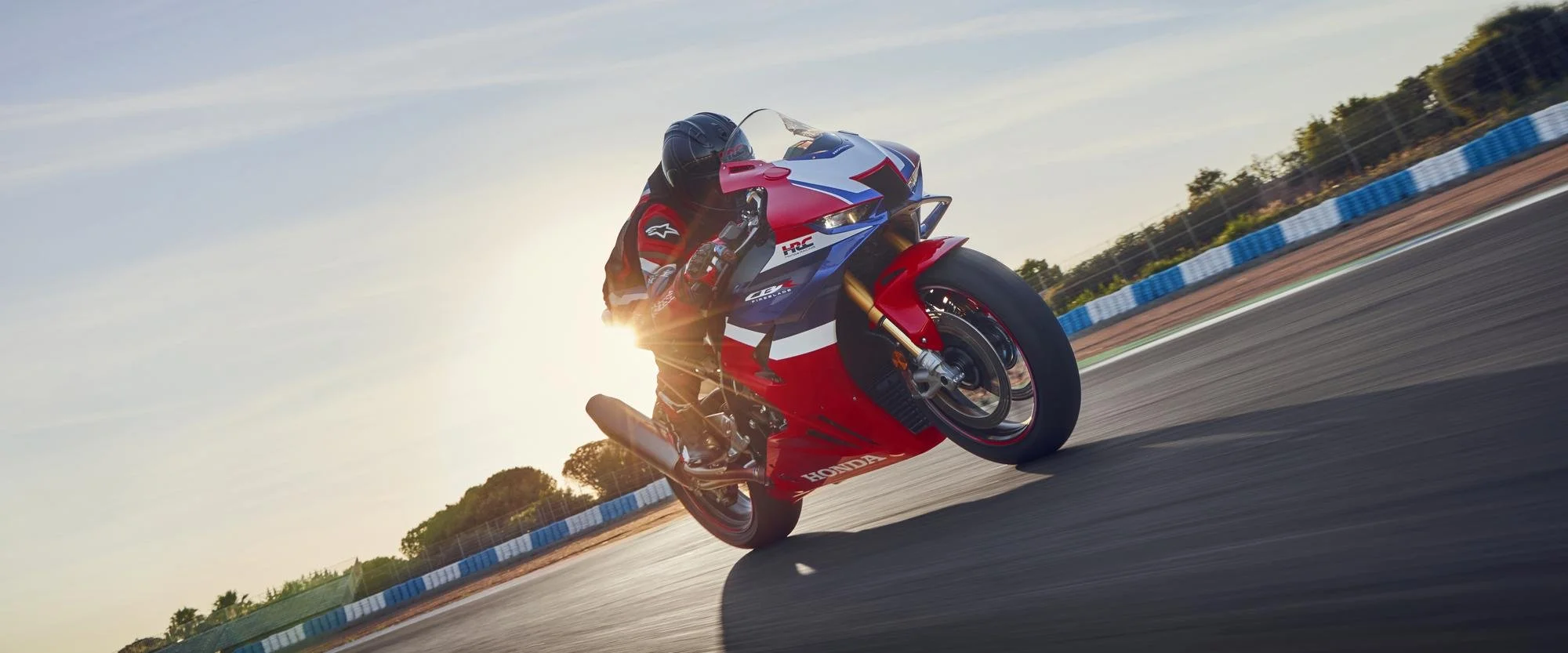 A motorcyclist leaning into a turn on a race track during sunset, riding a red, blue, and white Honda motorcycle.