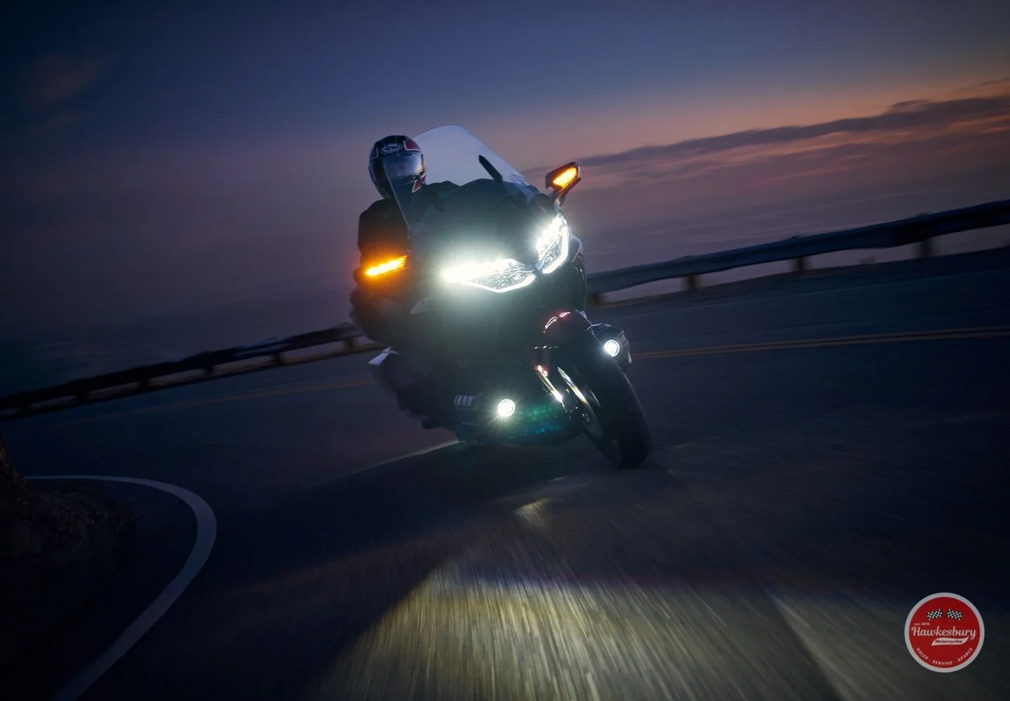 A motorcyclist riding on a curving road at dusk with the motorcycle's bright headlights and orange turn signals on, and the sky showing a sunset horizon.