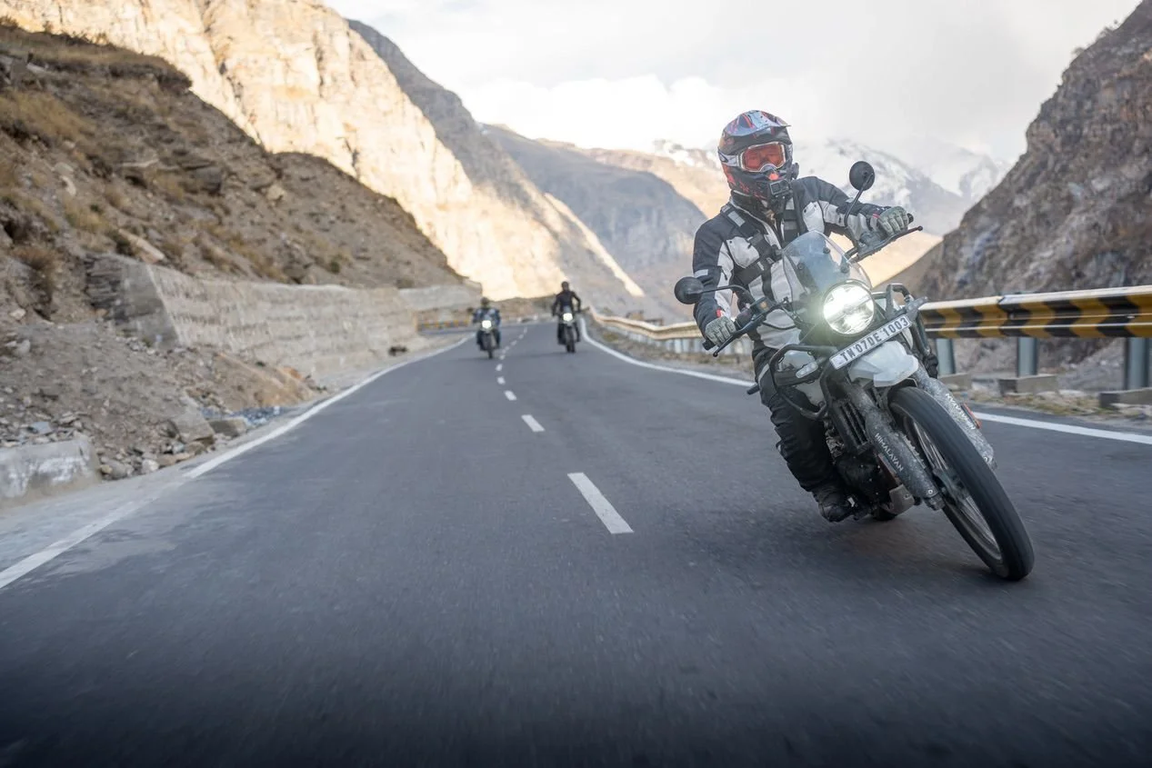 Motorcycle rider wearing a helmet and riding gear leaning into a curve on a mountain road with two other motorcyclists in the background, surrounded by rocky mountains.