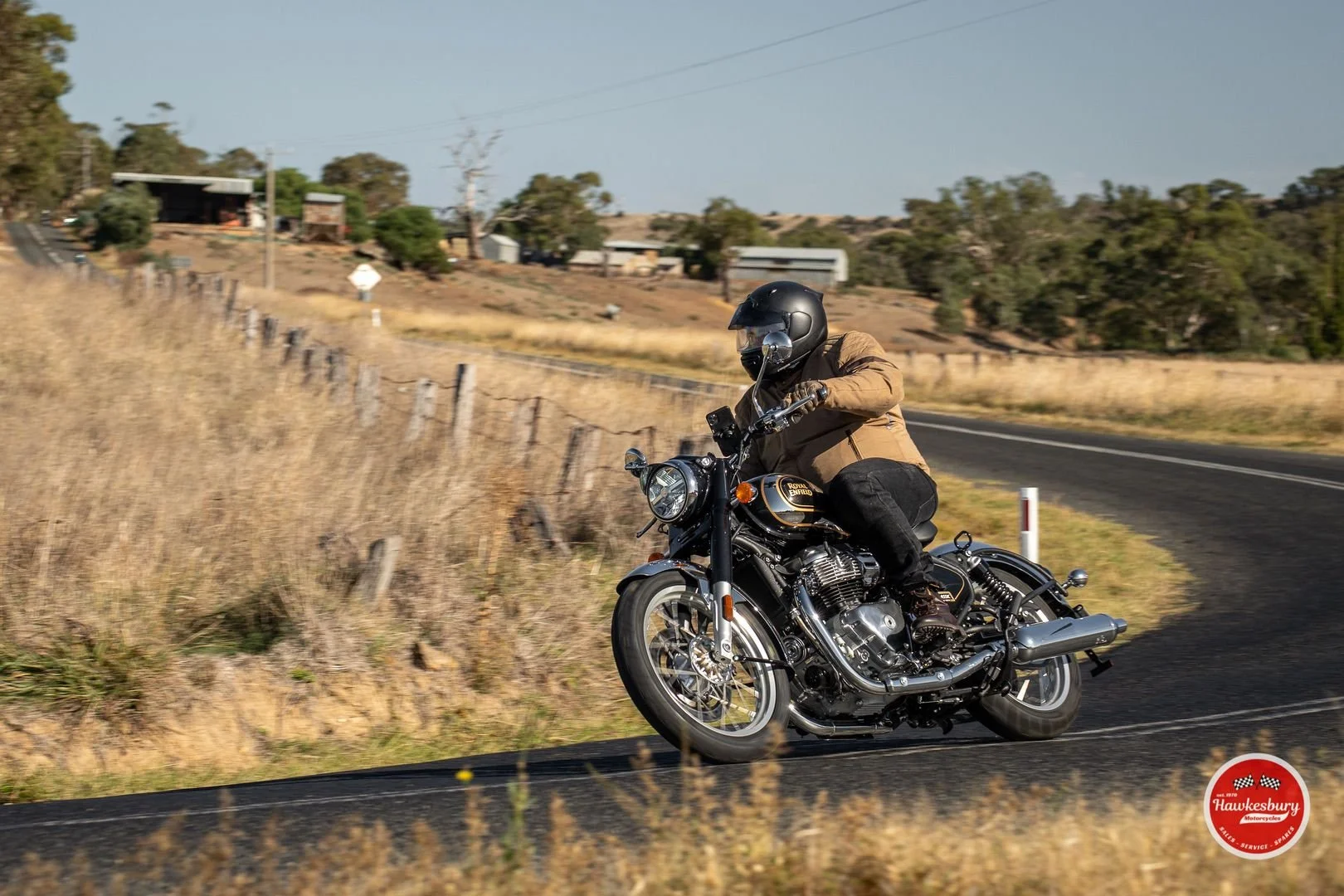 A motorcyclist riding a black Royal Enfield motorcycle on a curved rural road with fields, trees, and houses in the background.