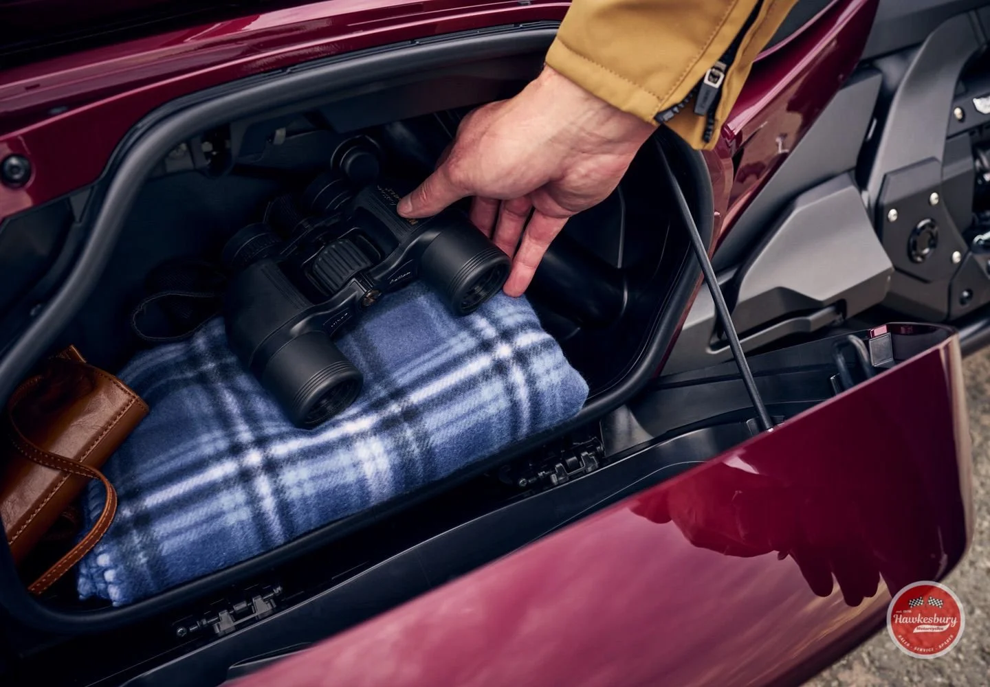 A person opening the front trunk of a red motorcycle, revealing a pair of binoculars and a blue plaid blanket inside.