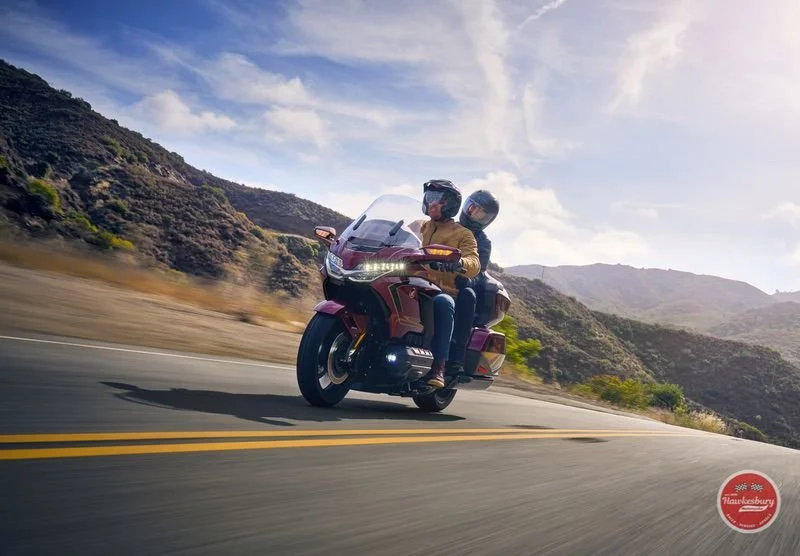 Two people riding a Honda Goldwing red touring motorcycle on a scenic mountain road under a partly cloudy sky.