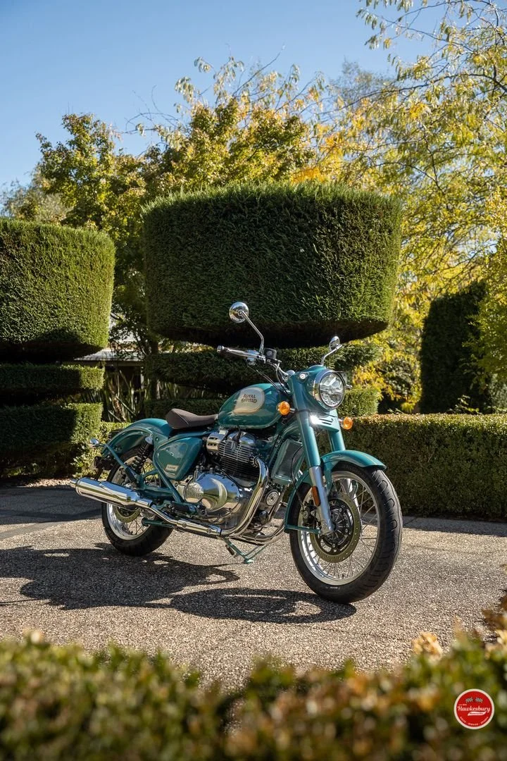 A teal Royal Enfield motorcycle parked outdoors on a paved surface, with neatly trimmed bushes and trees showing autumn foliage in the background.