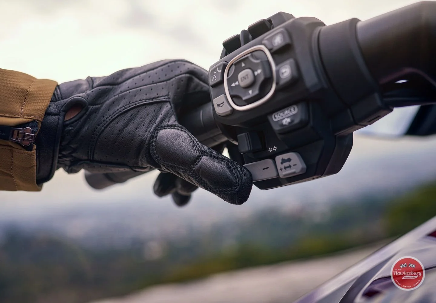Person wearing a black leather riding glove gripping a motorcycle handlebar with control buttons, with a blurred background and a red logo in the lower right corner.