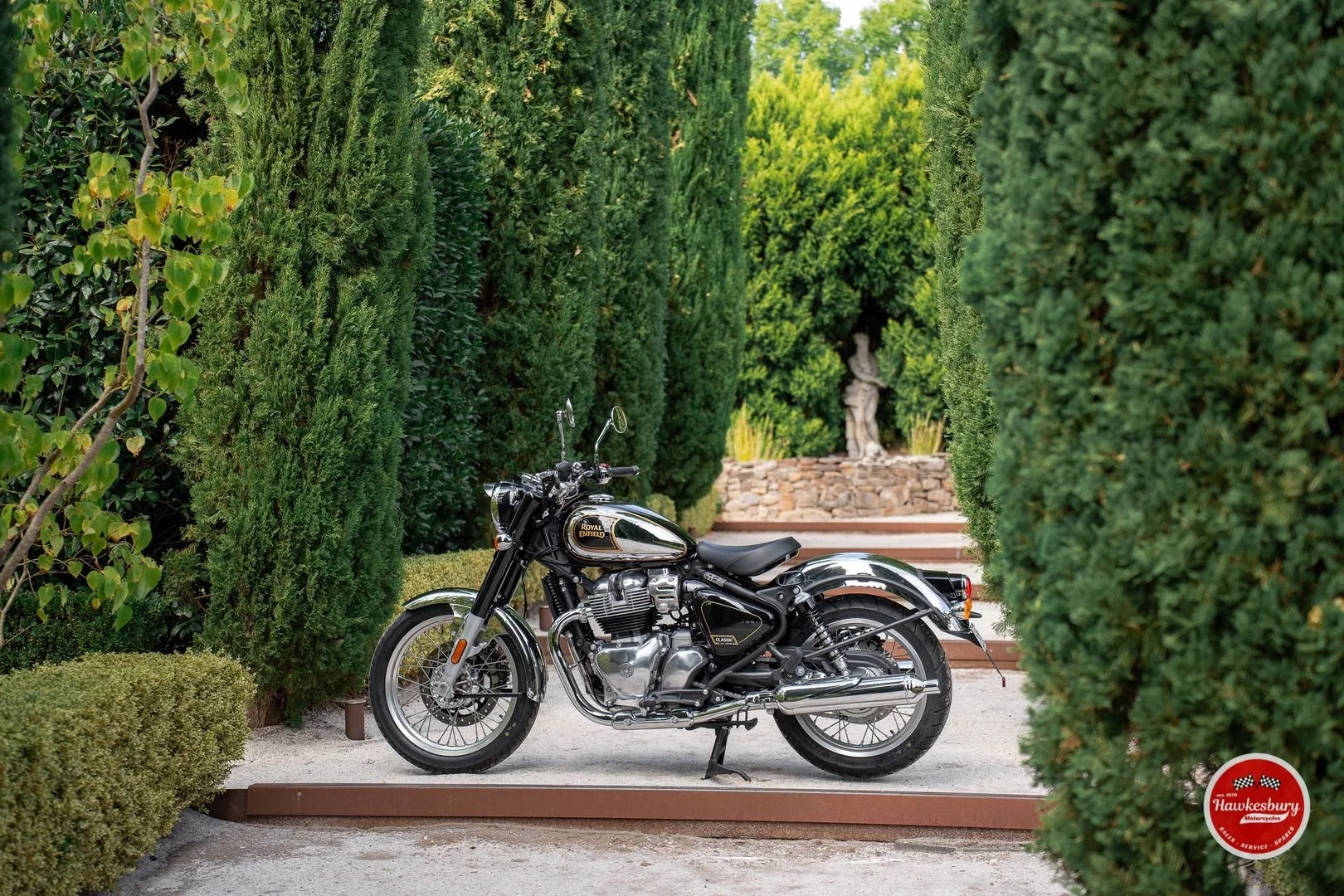 A black and chrome Royal Enfield motorcycle parked on a gravel path, surrounded by tall green hedges and trees, with a stone statue in the background.