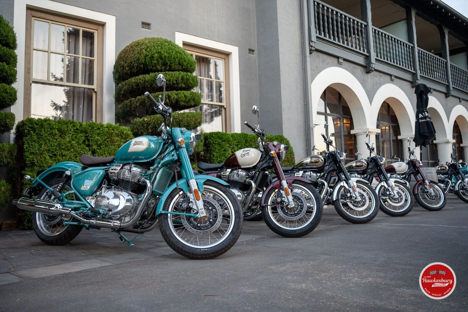 A row of vintage Royal Enfield motorcycles parked on the street outside a building with arched windows and a balcony, with neatly trimmed bushes in the background.